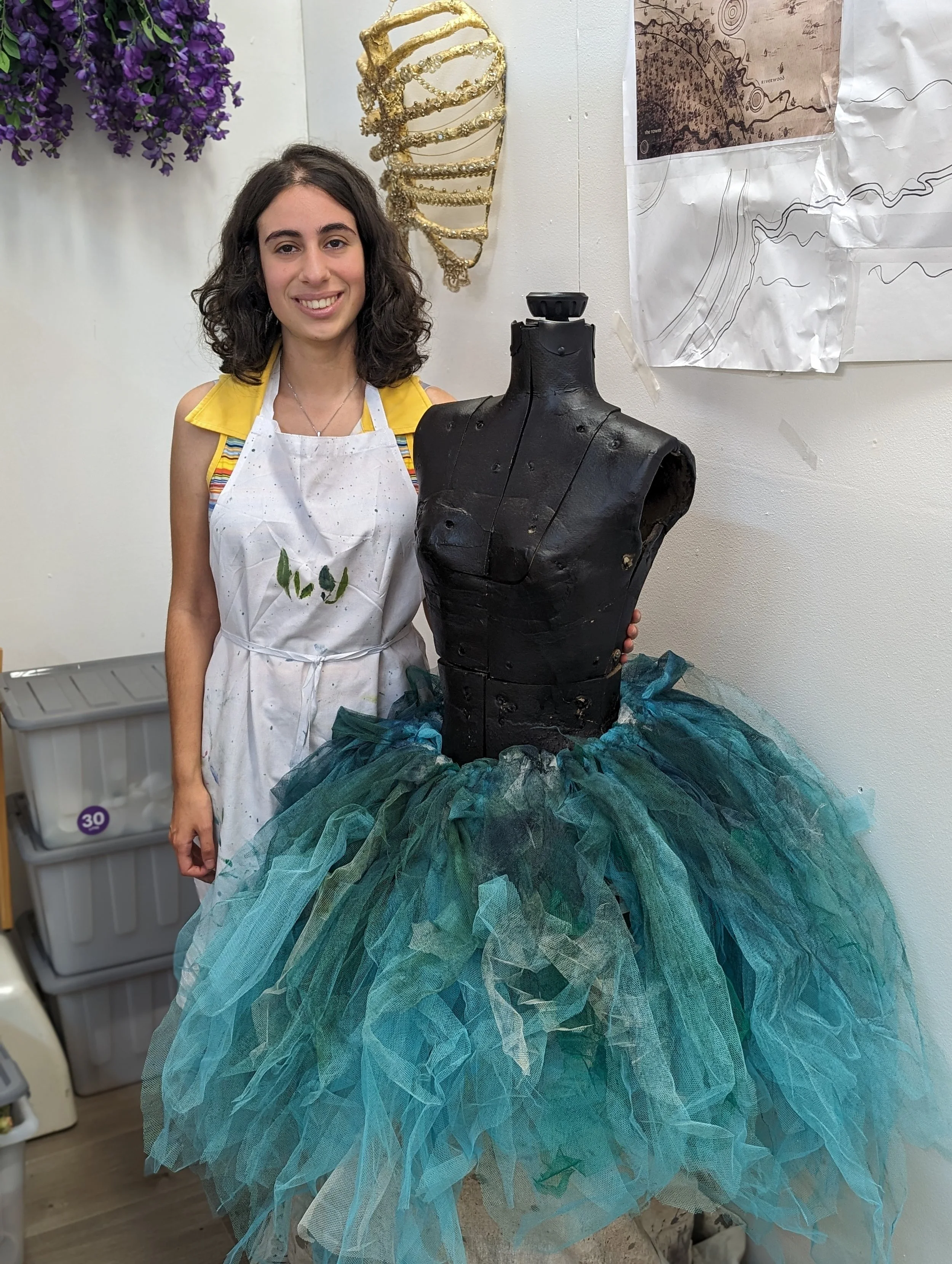 A woman with curly dark hair smiling while posing next to a black dress form decorated with blue and green tulle fabric, in an art or fashion studio with wall art and a gold sculpture in the background.