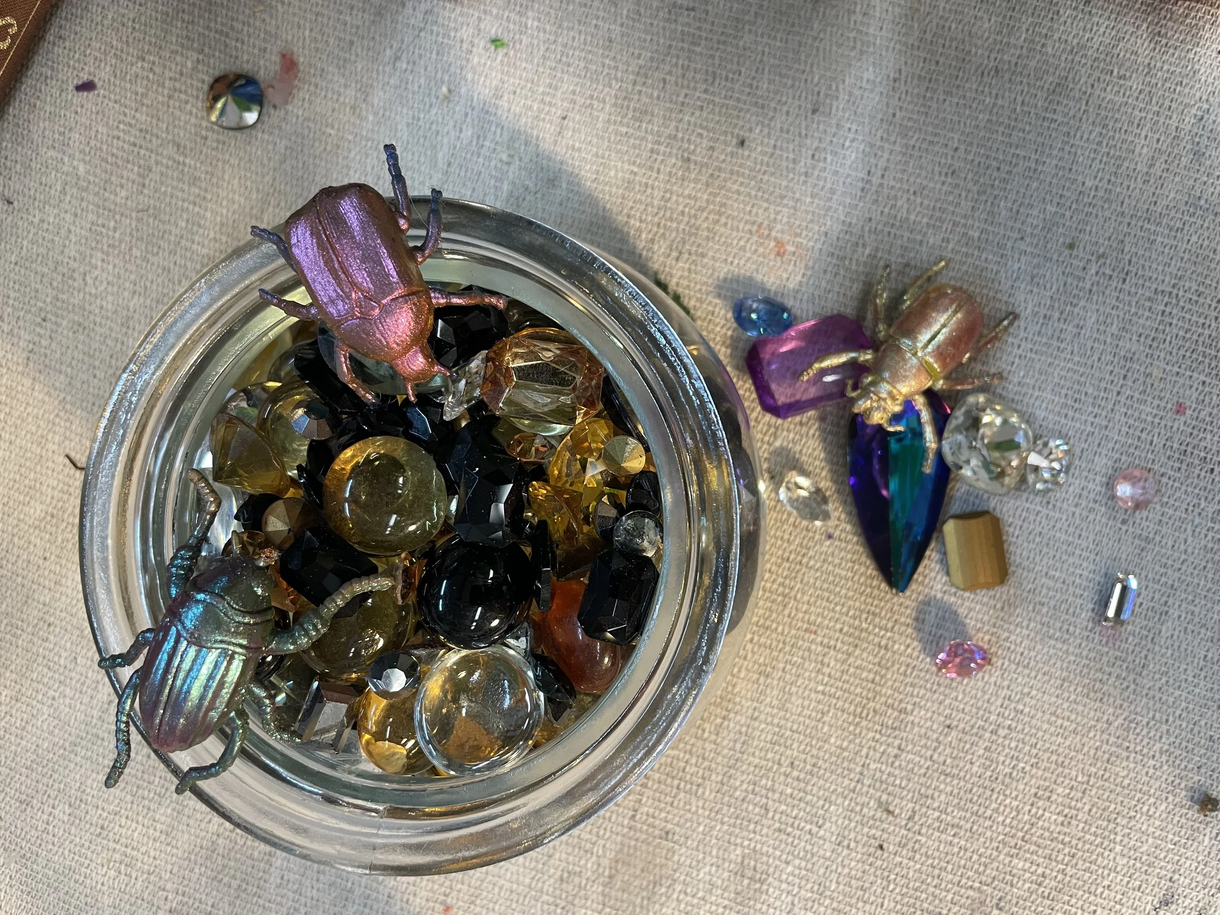 A container filled with various colorful gemstones and crystals, with a decorative beetle ornament on top, placed on a fabric surface with scattered small gems.