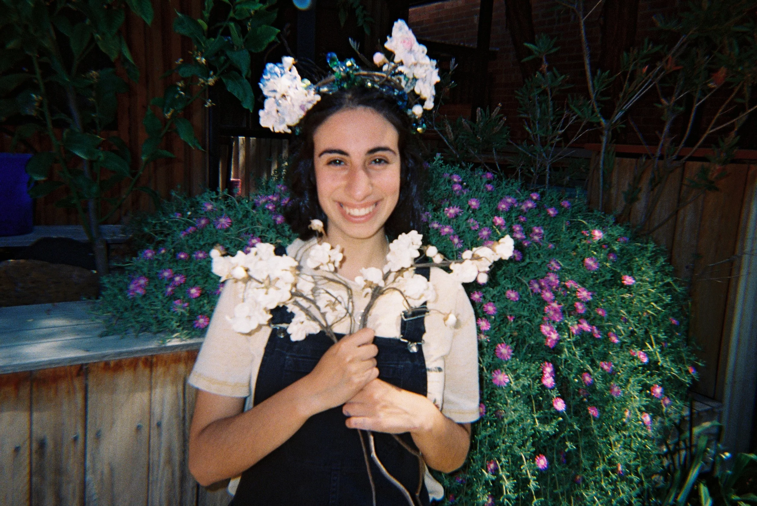 A young woman with dark curly hair wearing a floral crown and overalls, smiling while holding a branch of white flowers, standing in front of a garden with purple and green plants.