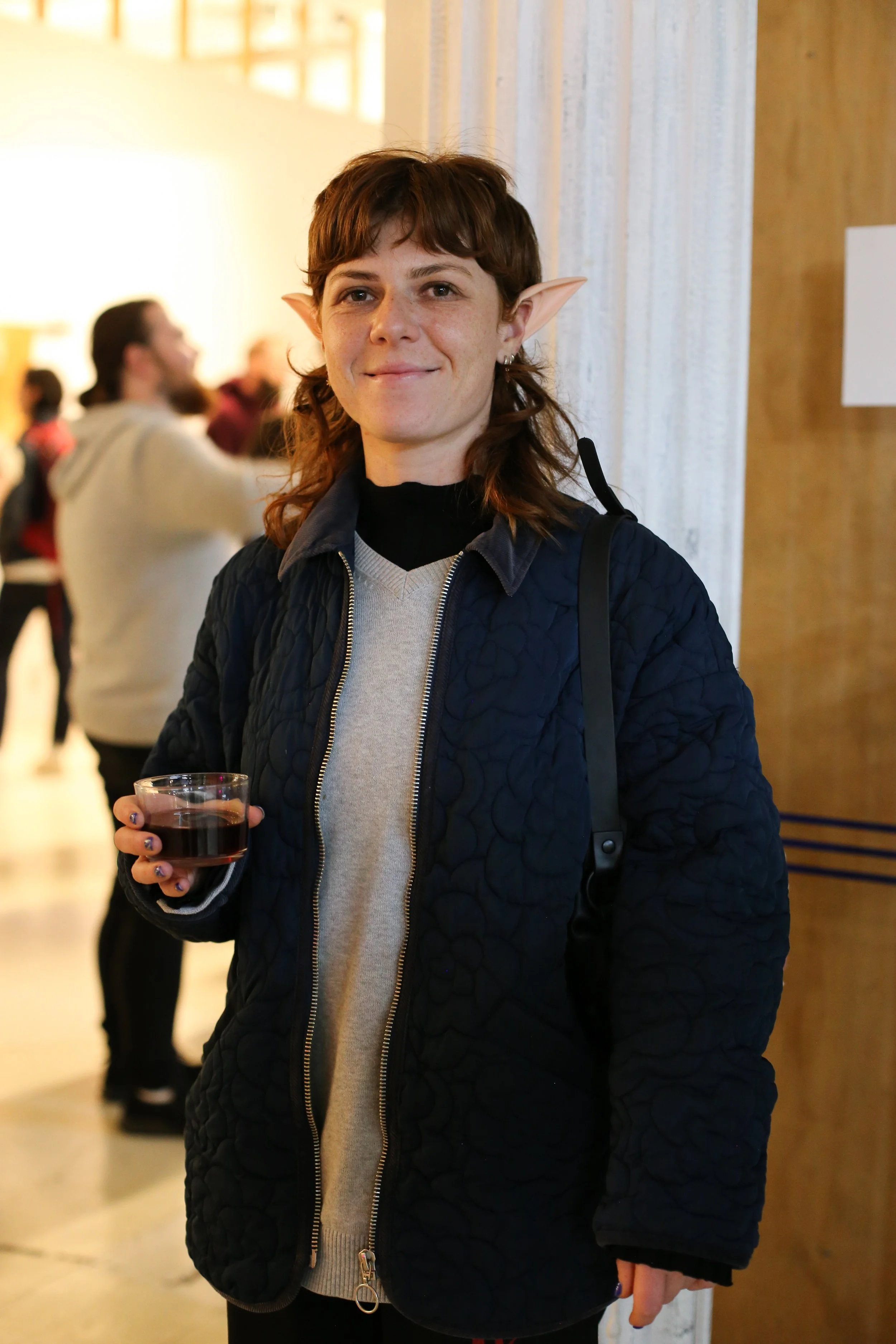 A person with elf-like ears, short brown hair, and light skin, smiling and holding a glass of red wine, standing indoors in a crowded space.