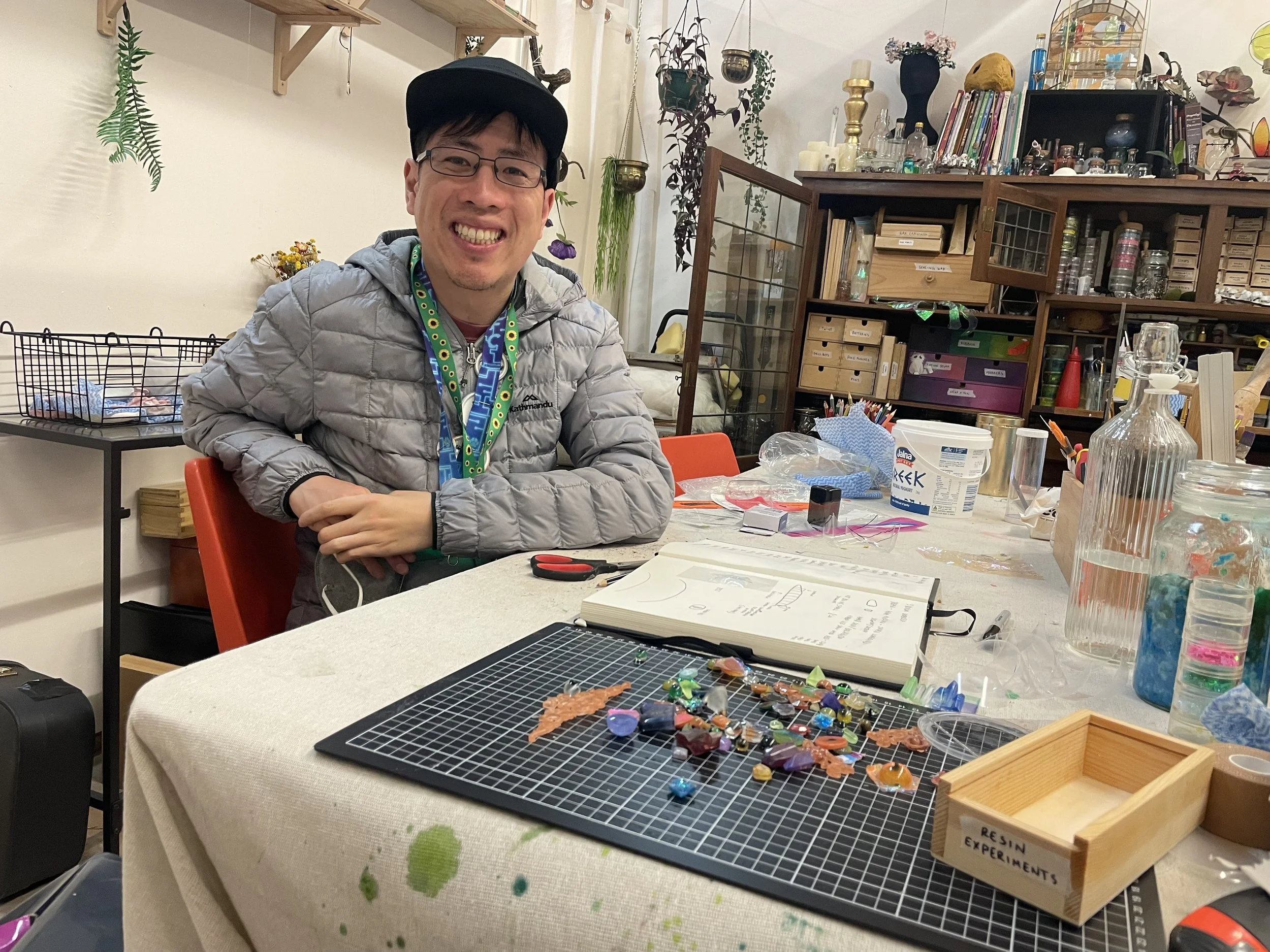 A man wearing glasses, a cap, and a grey jacket smiling at a crafting or jewelry-making workspace with various colorful stones, tools, and supplies on a table.
