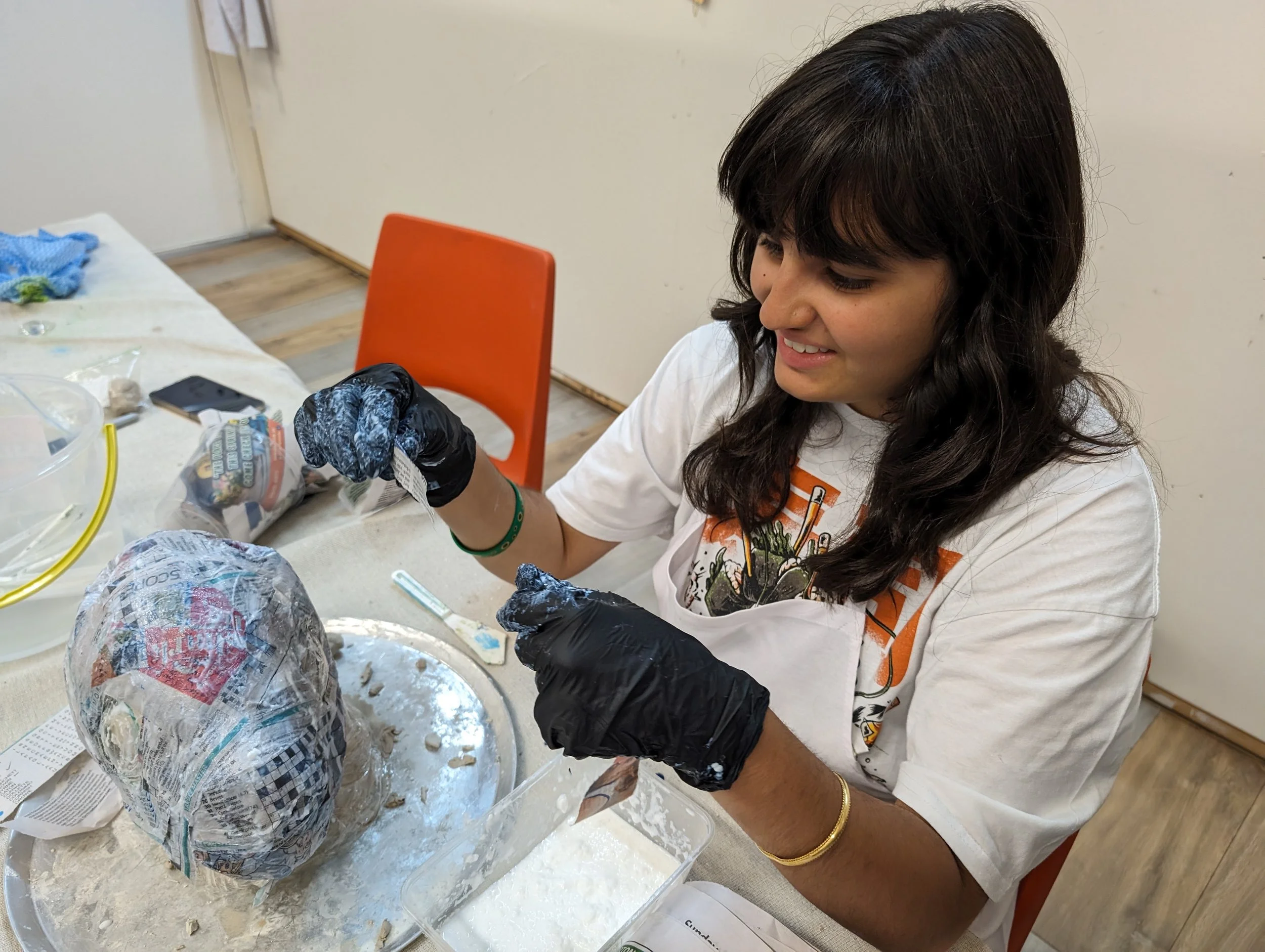 A woman with dark hair and a nose piercing is sitting at a table, smiling, as she works on a papier-mâché project involving newspaper and spray paint. There are various crafting supplies on the table.