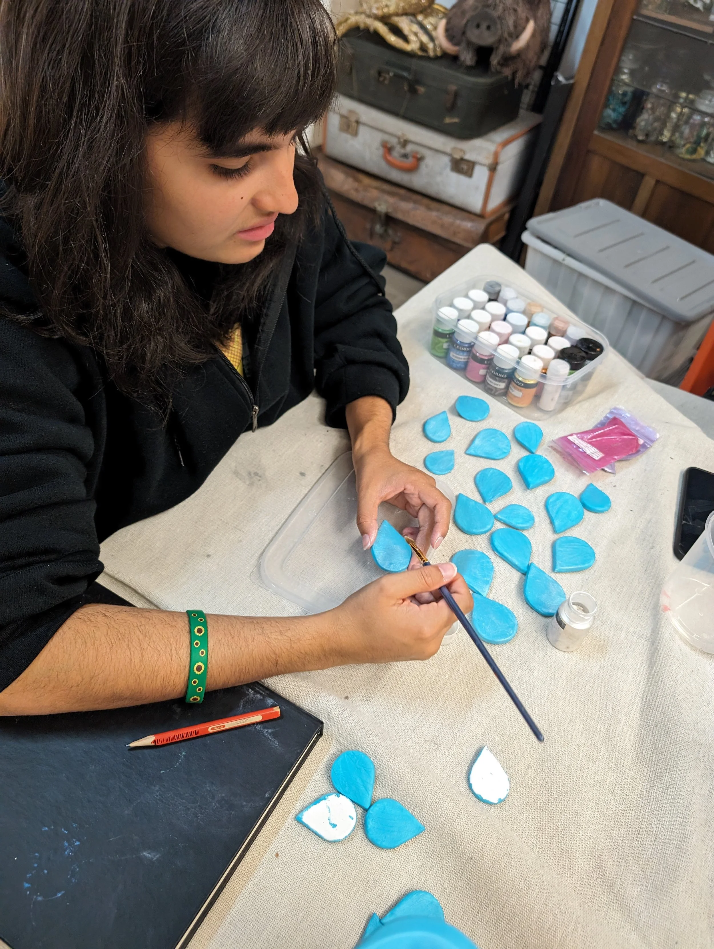 Person working on painting blue scales with a small paintbrush at a table, containers of paint nearby, in a room with vintage storage trunks and shelves in the background.