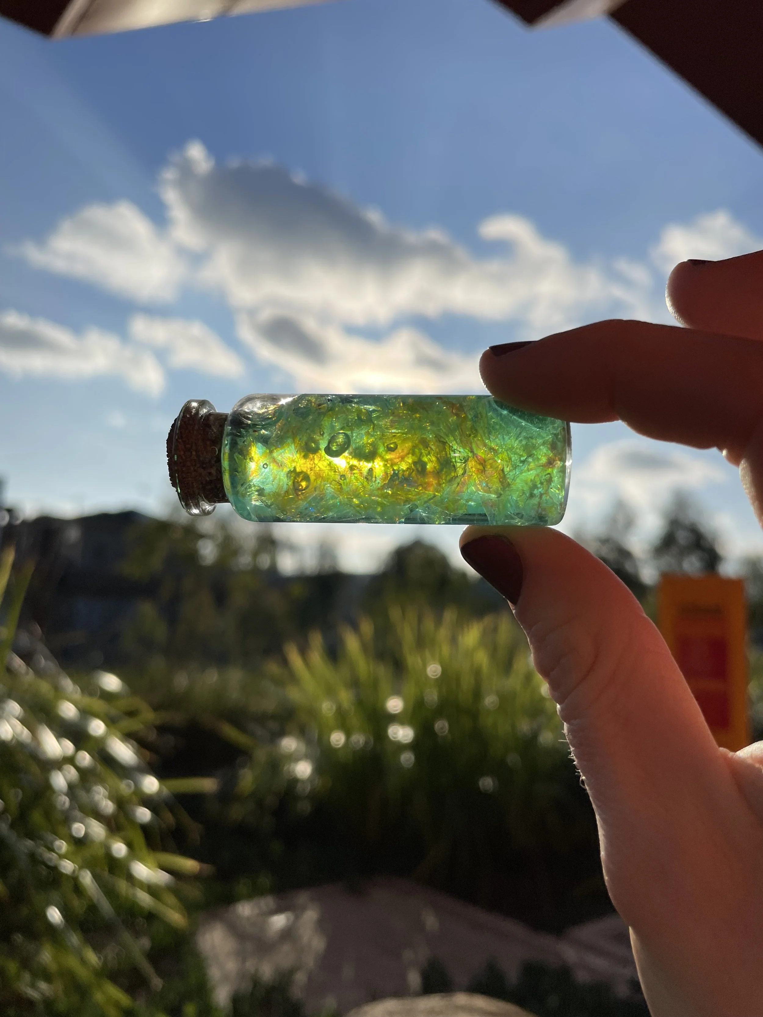 A small glass vial with yellow and green swirling patterns and bubbles inside, being held by a person's fingers against a blue sky with scattered clouds.