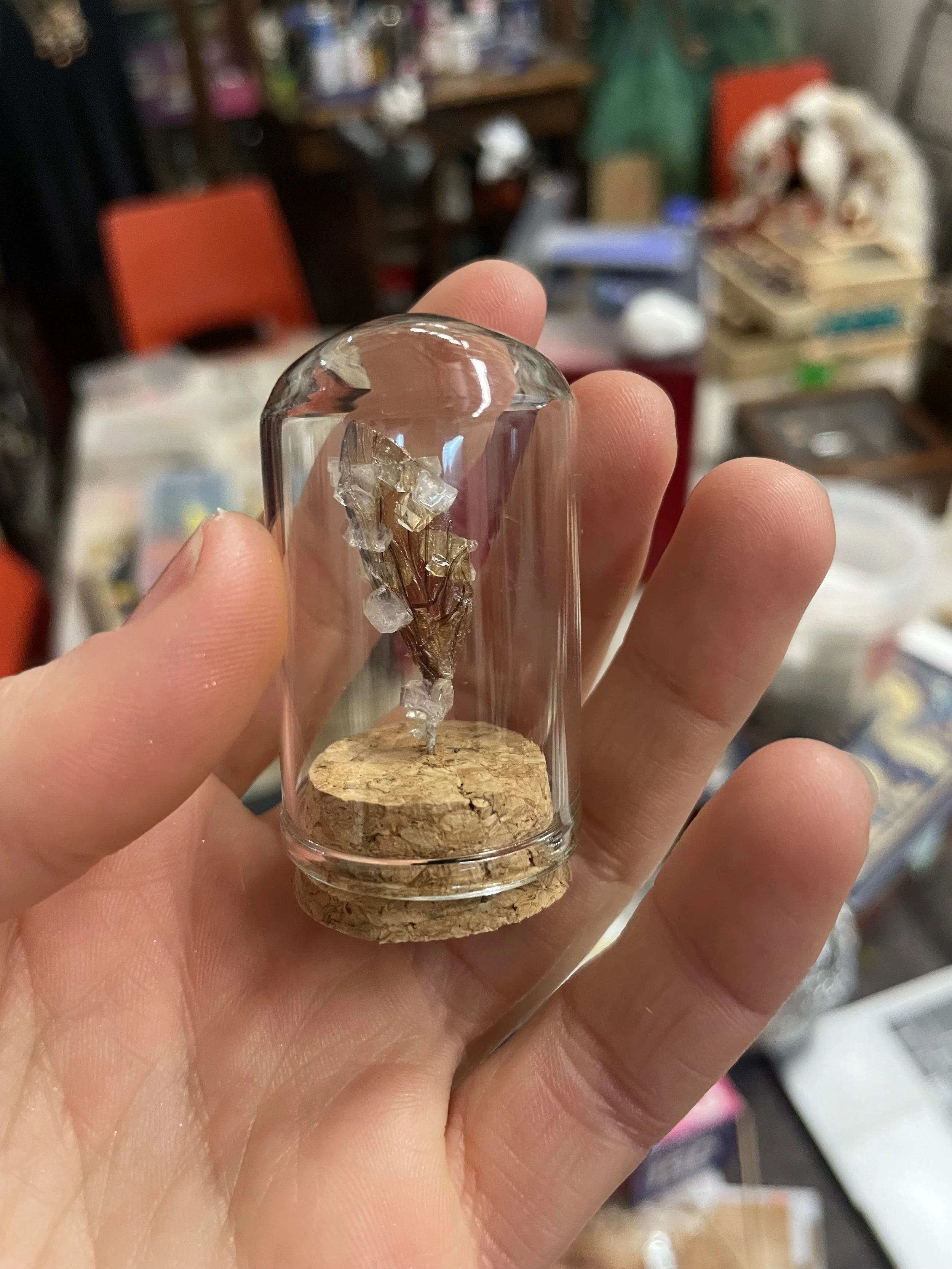 A hand holding a small glass display case with a cork base, containing a gemstone or mineral specimen with transparent and shiny crystals.