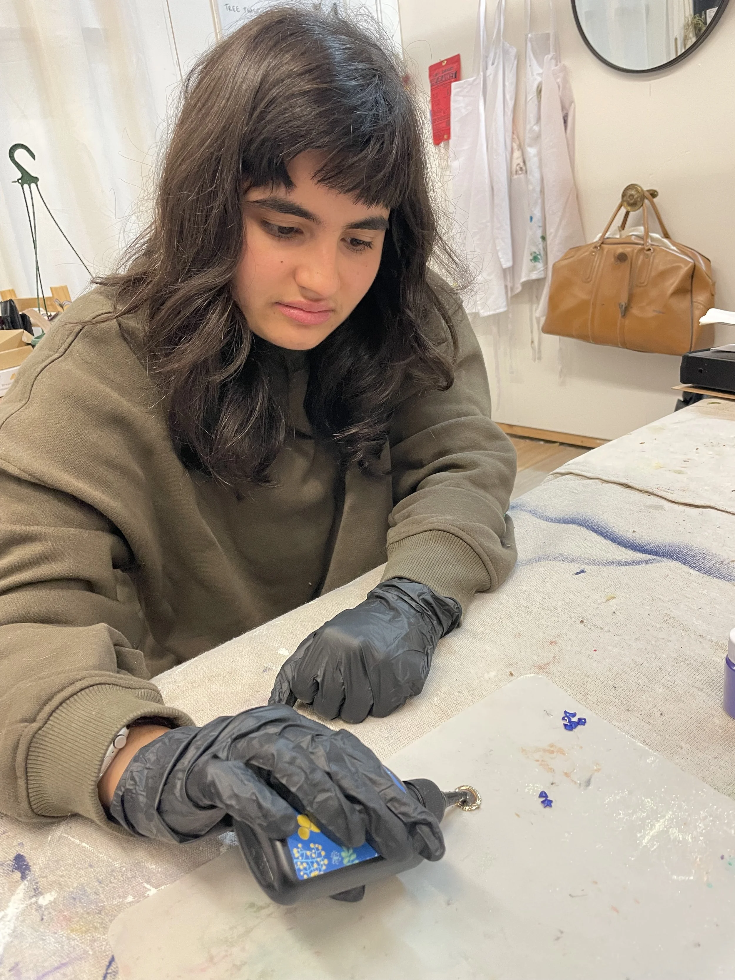 A young woman with dark hair, wearing a brown sweatshirt, is working on a craft project at a table. She is wearing black gloves and holding a tool with a yellow tip. The table has scattered small blue items and some paint stains. In the background, t