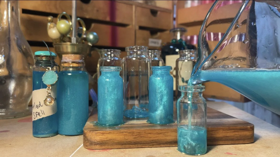 Various glass bottles filled with blue-colored liquid or crystals on a wooden surface, with one pouring the liquid into a small bottle, in a craft or display setting.
