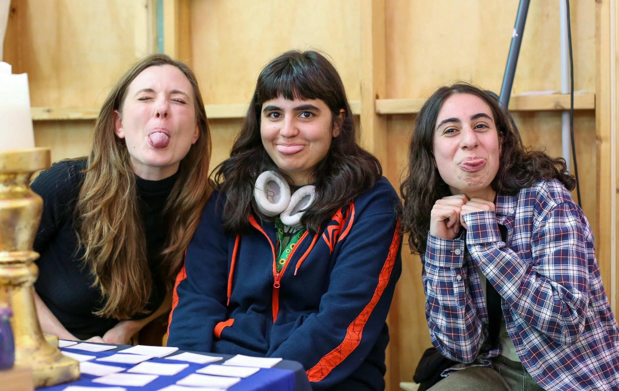 Three young women sitting at a table, making silly faces and sticking out their tongues, in a wooden-paneled room.