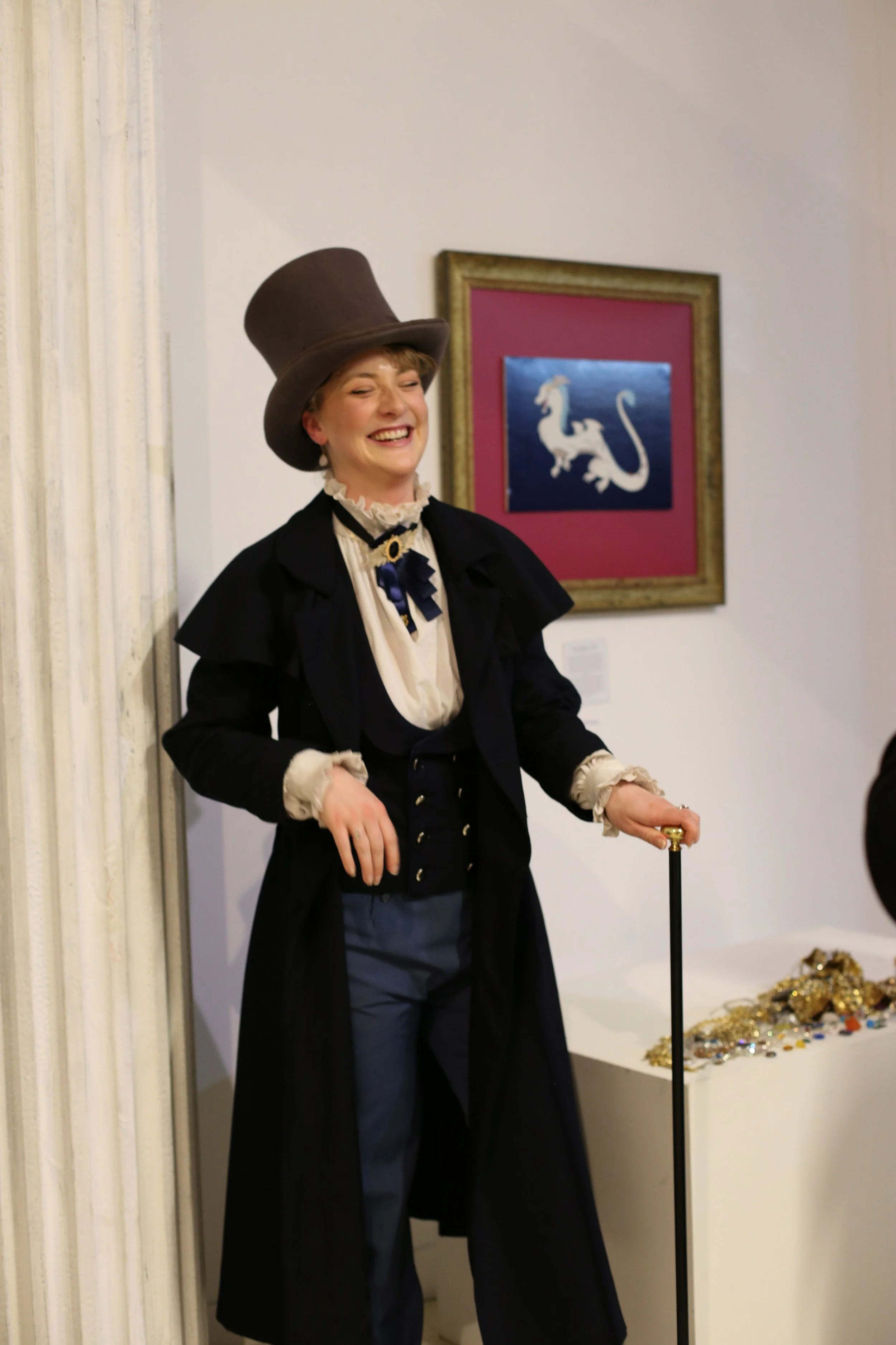 Woman dressed as a Victorian character, smiling, wearing a tall top hat, black coat, and vintage-style blouse, standing indoors next to a display of jewelry, with a framed dragon on the wall behind her.