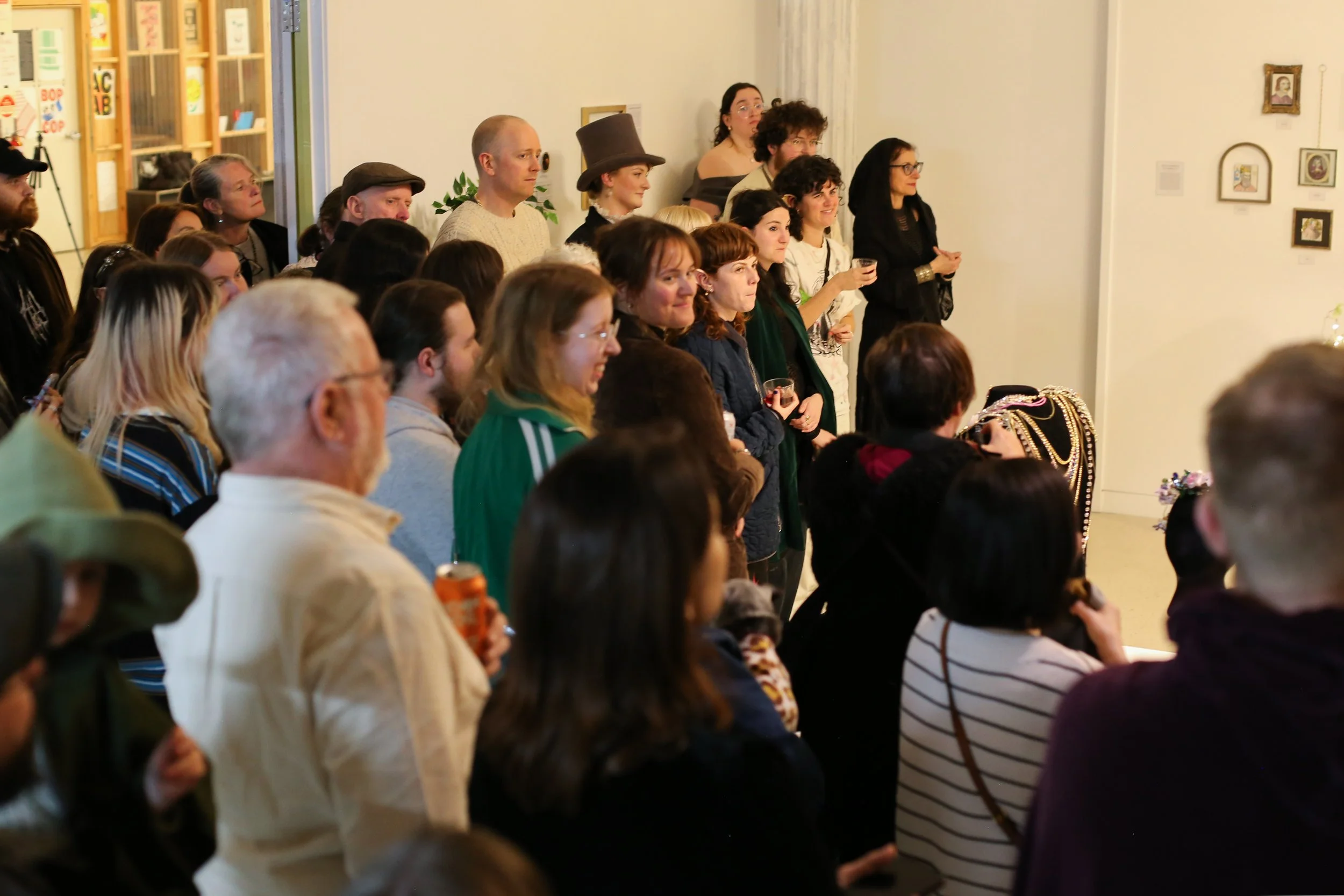 A group of people gathered in an art gallery, watching an artwork or event outside the frame. The crowd includes men and women of various ages, some smiling and holding drinks, with some wearing casual and costume-like attire, near a wall decorated w