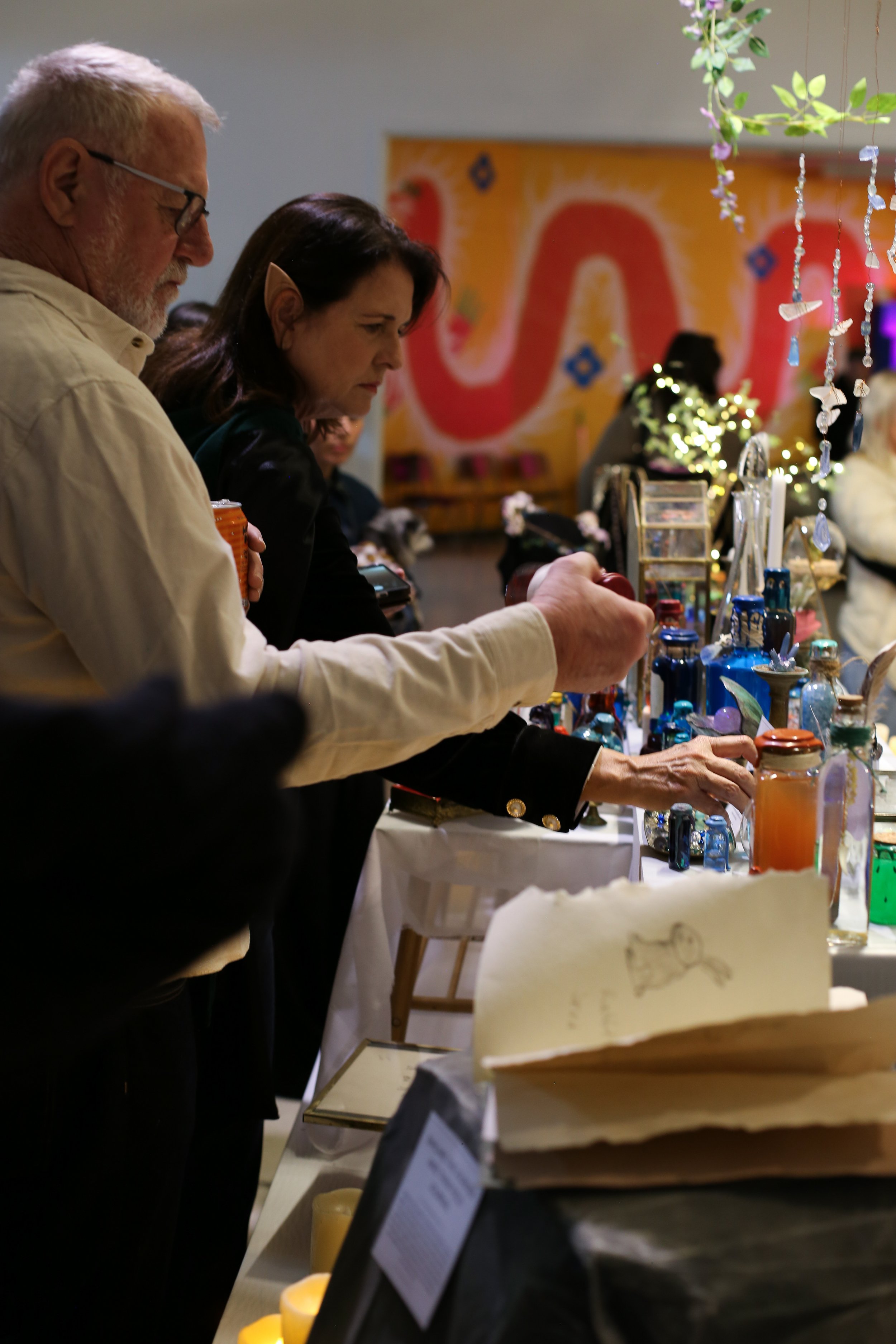 Two people, a man with gray hair and beard, and a woman with dark hair and elf-like ear prosthetic, are looking at items on display at. The display includes colorful jars, bottles, and decorative hanging crystals, with a large abstract painting in th