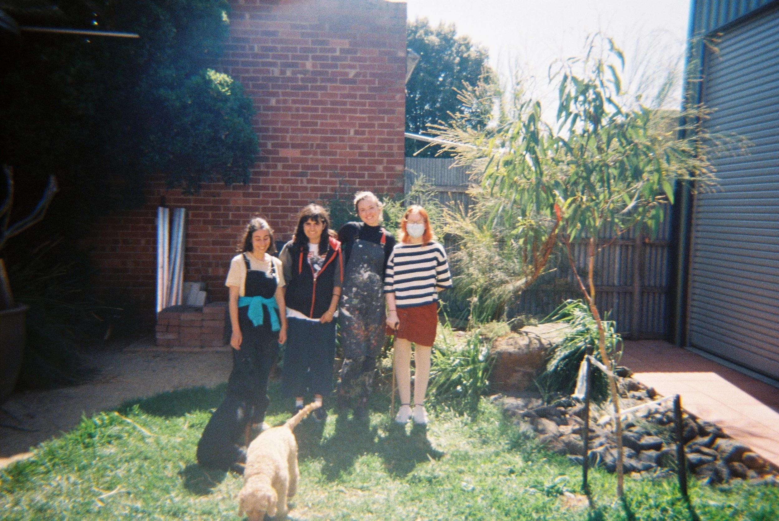 Four women standing outdoors in a garden near a brick wall and a tree. One woman is wearing a face mask, all are smiling, and a dog is lying on the grass in front of them.