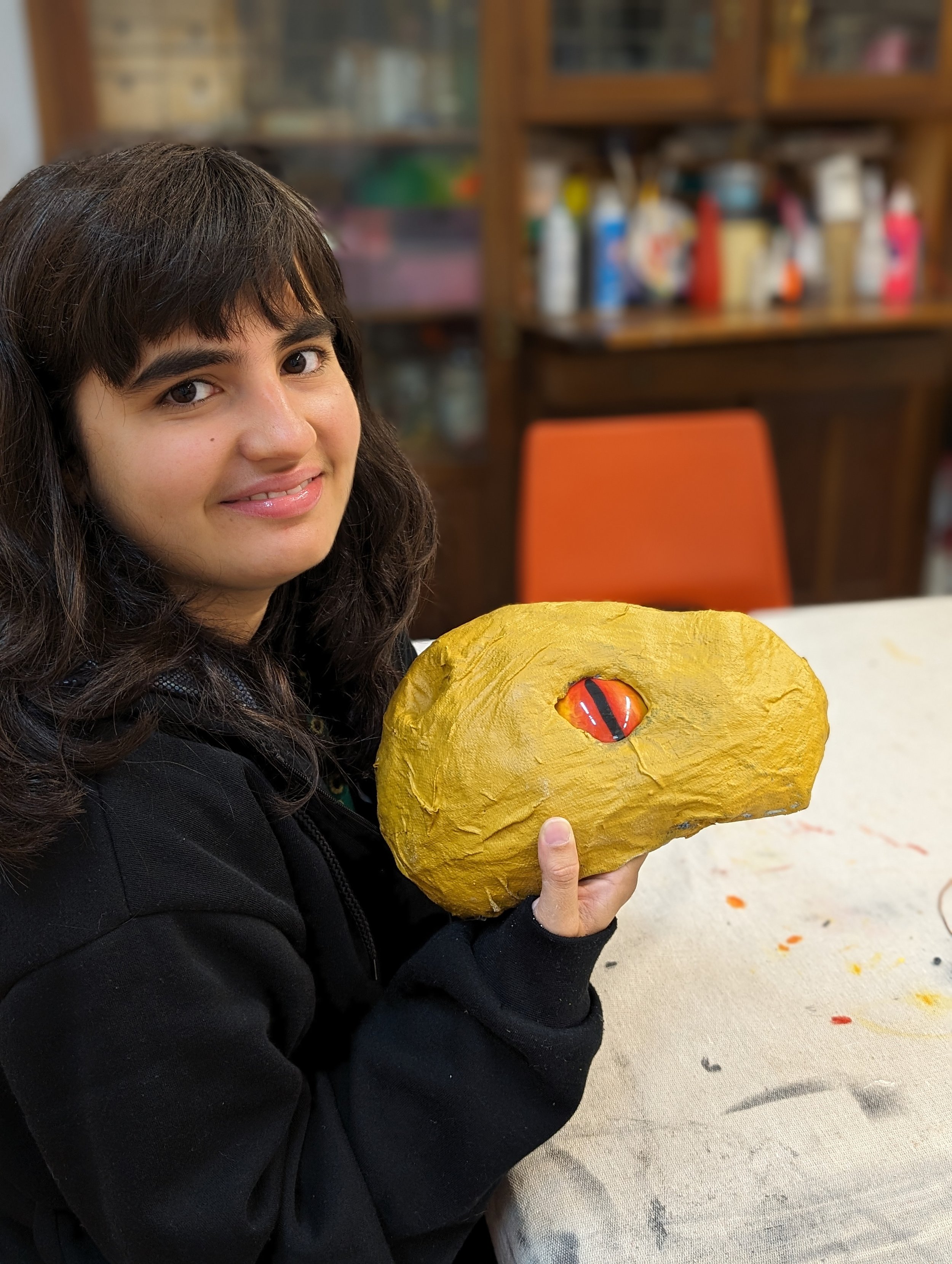 A young girl with dark brown hair and a black jacket holding a craft project of a dragons head, sitting at a table with paint splatters, in a room with wooden cabinets in the background.