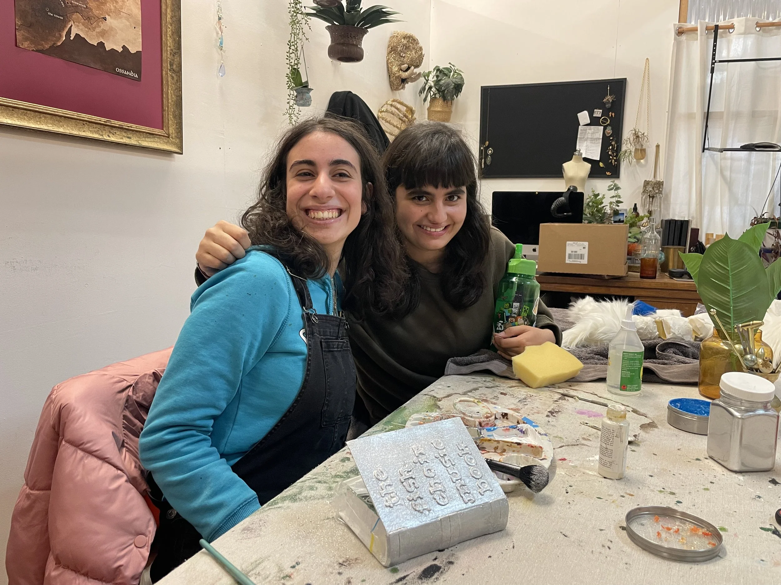Two young women are sitting at a table, smiling at the camera in an art studio. The table is covered with art supplies and decorated with painted plaques and painted decoration items. The woman on the left is wearing a turquoise hoodie and a pink puf