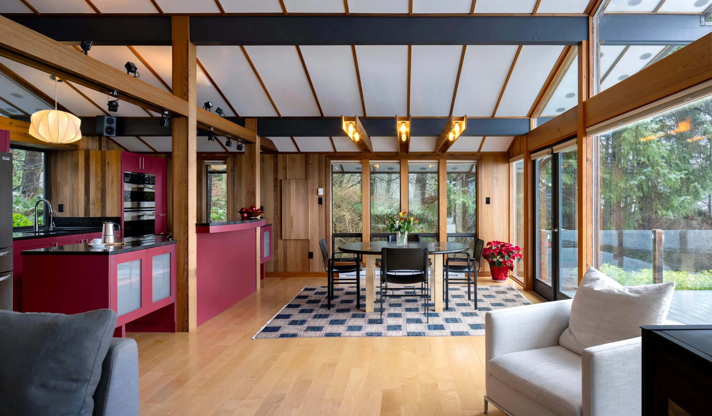 Interior of a modern living and dining area with wooden walls and ceiling beams, large windows showcasing a forest view, a round dining table with chairs, a rug underneath, and a white armchair in the foreground.