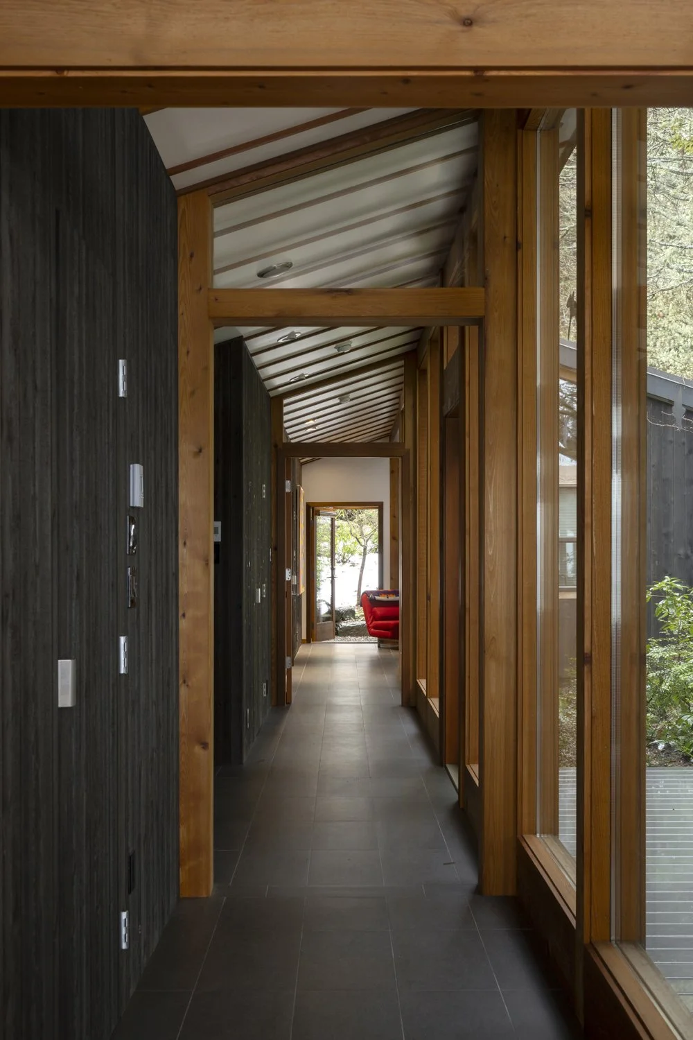 Wooden corridor with large windows on the right side, leading to a sunlit room with a red chair and outdoor greenery visible through the door.