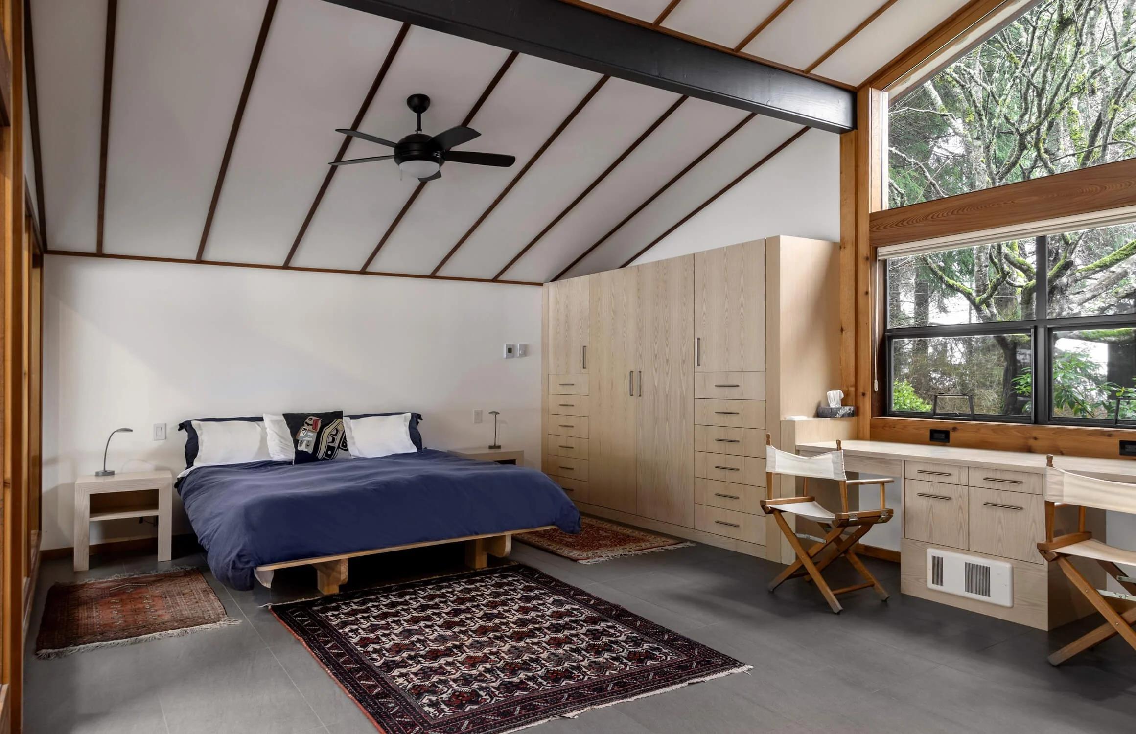Bedroom with dark wood ceiling, white walls, large window showing trees outside, bed with dark blue bedding, two bedside tables, built-in wardrobe, and natural wood furniture.