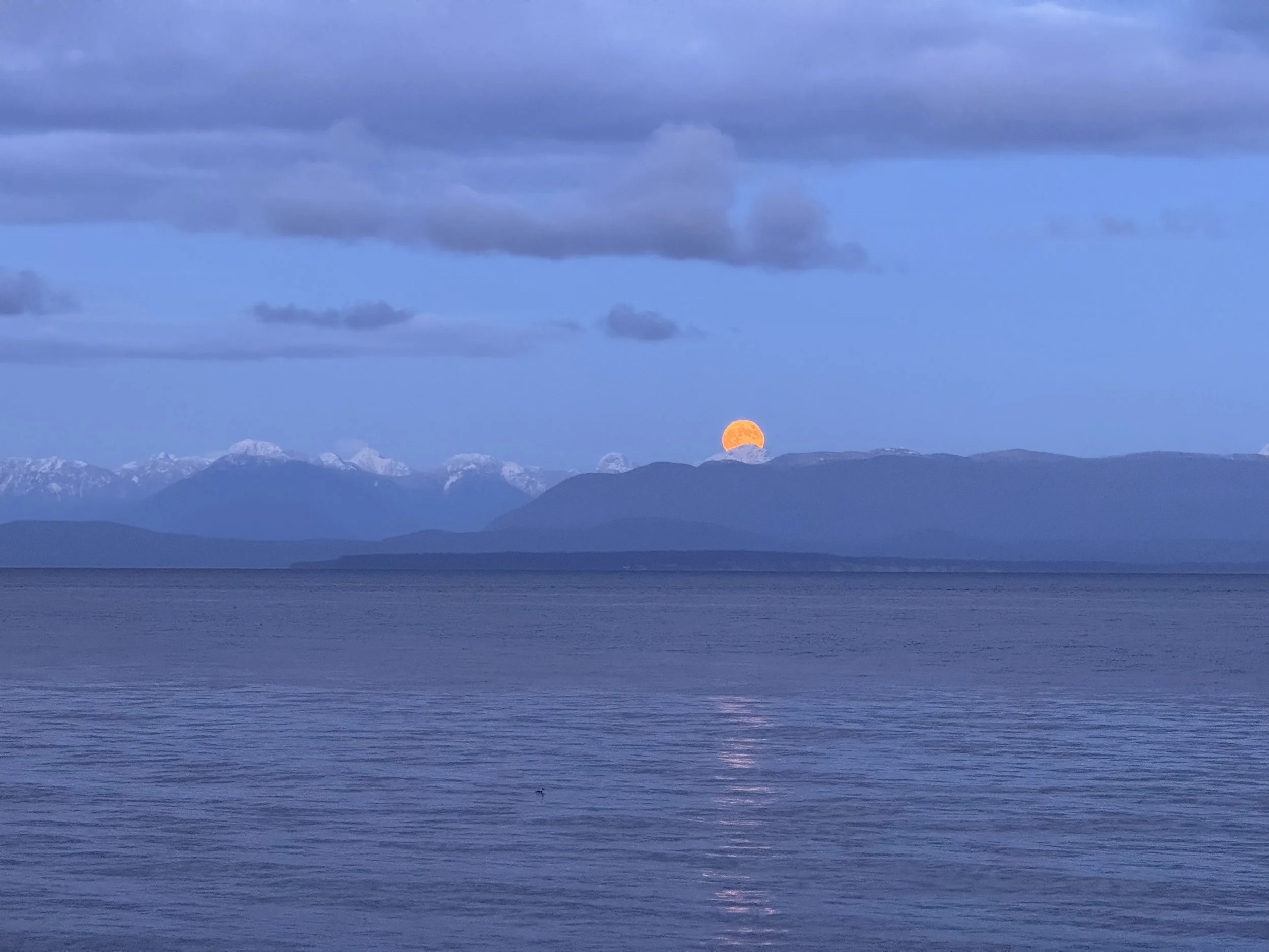 Full moon rising over a mountain range at dusk, with snow-capped peaks in the background and a calm body of water in the foreground reflecting the moonlight.