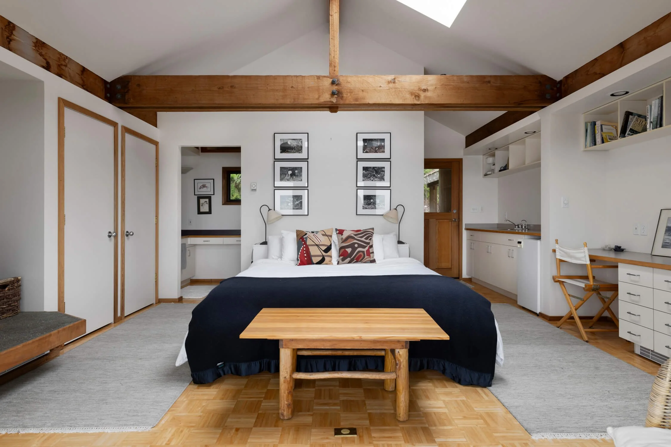 A cozy bedroom with white walls and wood accents. Central bed with white bedding, black blanket, and decorative pillows. Framed black and white photos above the bed. Wooden bench at the foot of the bed. Small kitchen area on the right, work desk with chair. Open closet doors on the left and natural light coming through windows and skylight.