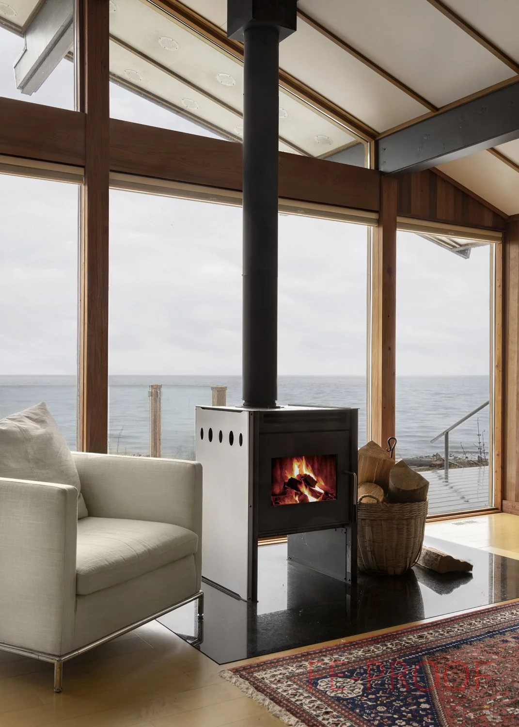 Living room with large windows overlooking the ocean, a white sofa, a wood stove with a fire burning, a basket of firewood, and a decorative carpet.