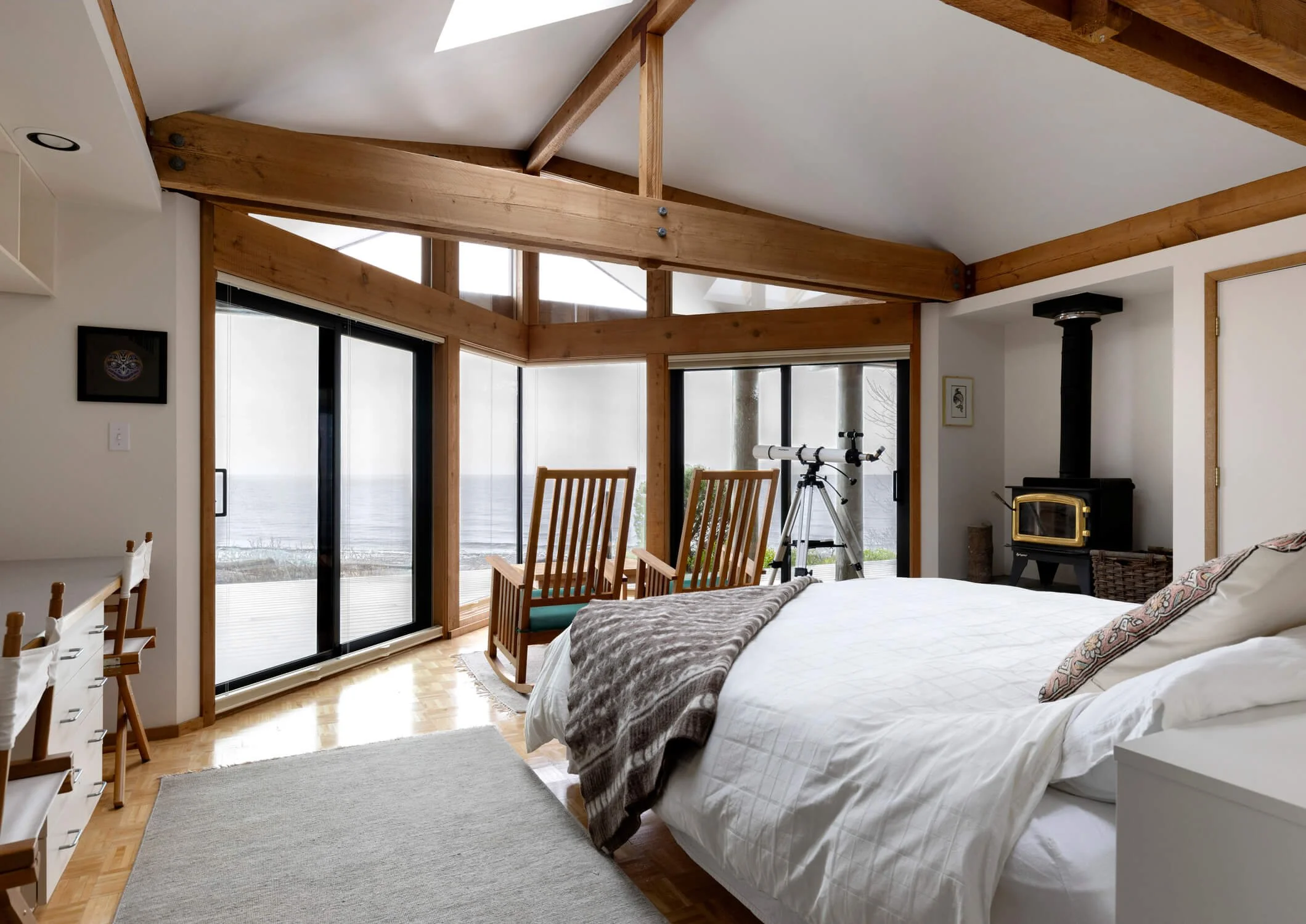 A bedroom with large windows and sliding glass doors showing an ocean view. The room has wooden beams, a white bed with a brown blanket, a telescope, a wood stove, and two wooden chairs facing the window.