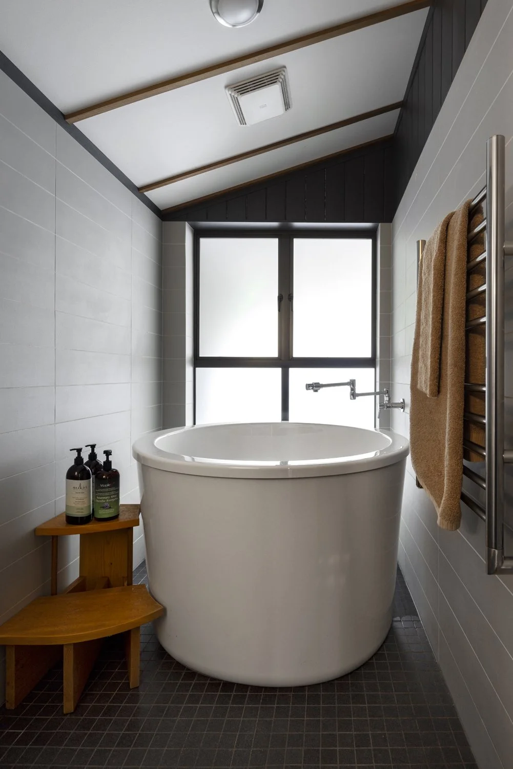 Modern bathroom with a free-standing bathtub near a large window, brown towels on a wall-mounted heated towel rail, and a small wooden side table holding soap bottles.