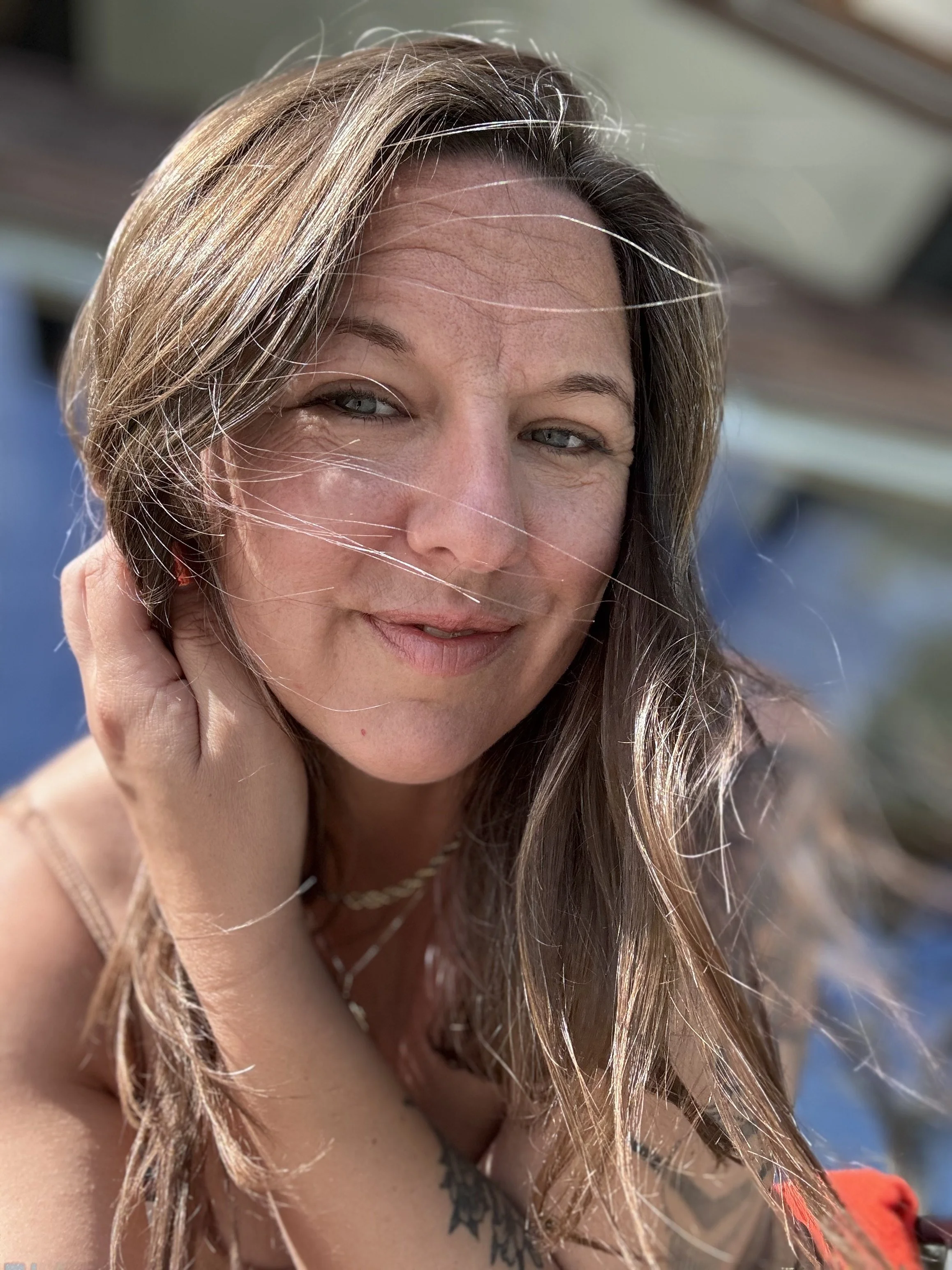 Close-up of a woman outdoors with long, light brown hair, smiling slightly, with wind blowing her hair, wearing a tank top and a necklace.