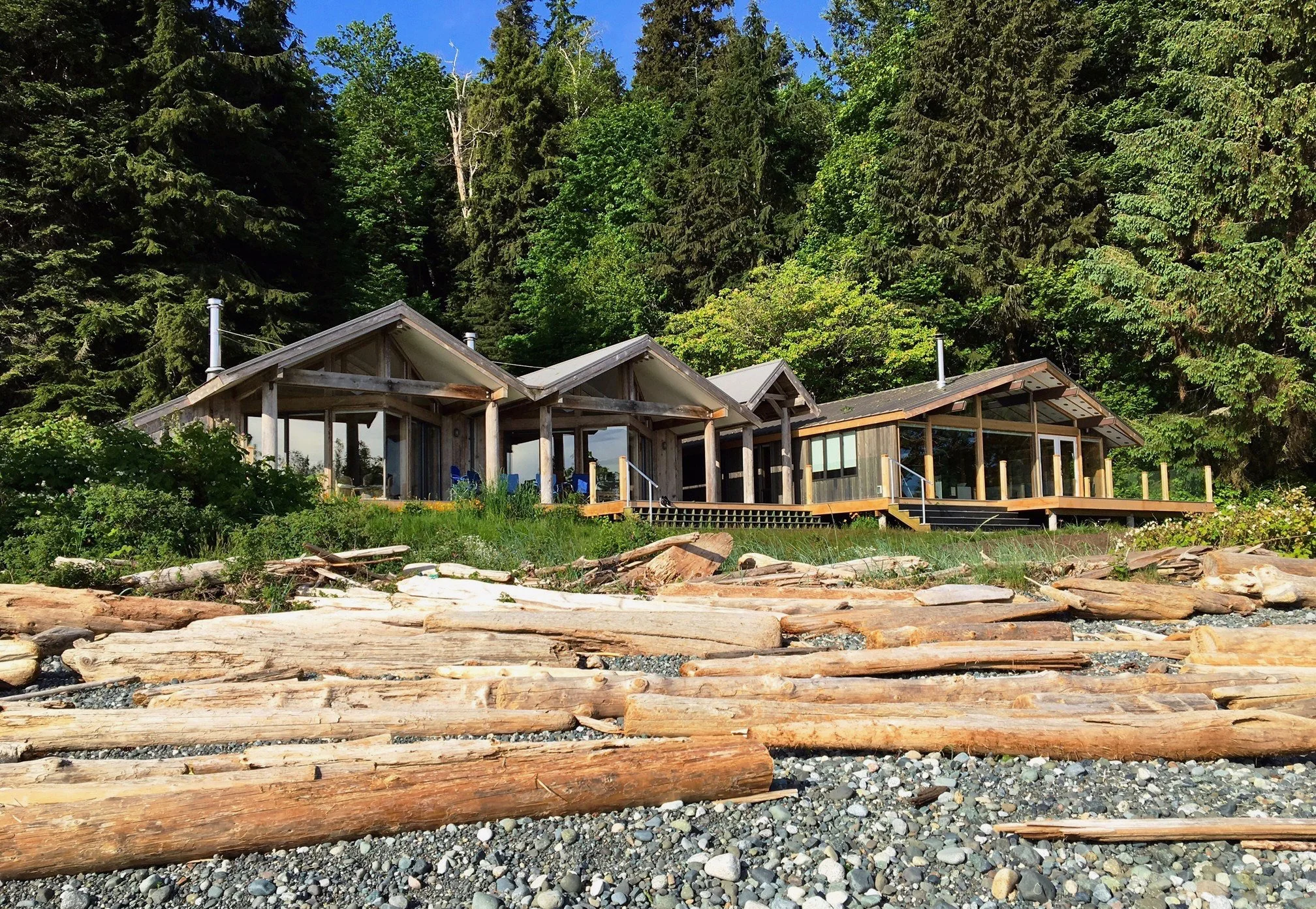 A modern wooden house built on a rocky beachfront with a forested backdrop, featuring large glass windows and a deck, under a clear blue sky.