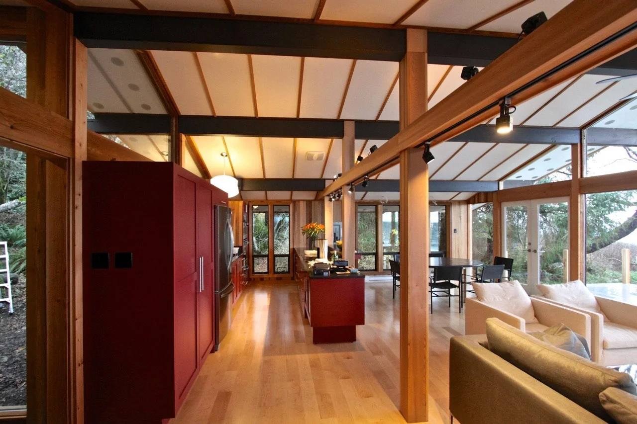 Interior view of a modern open-concept living space with a kitchen, dining area, and living room, featuring wooden beams, hardwood floors, and large windows overlooking a wooded outdoor area.