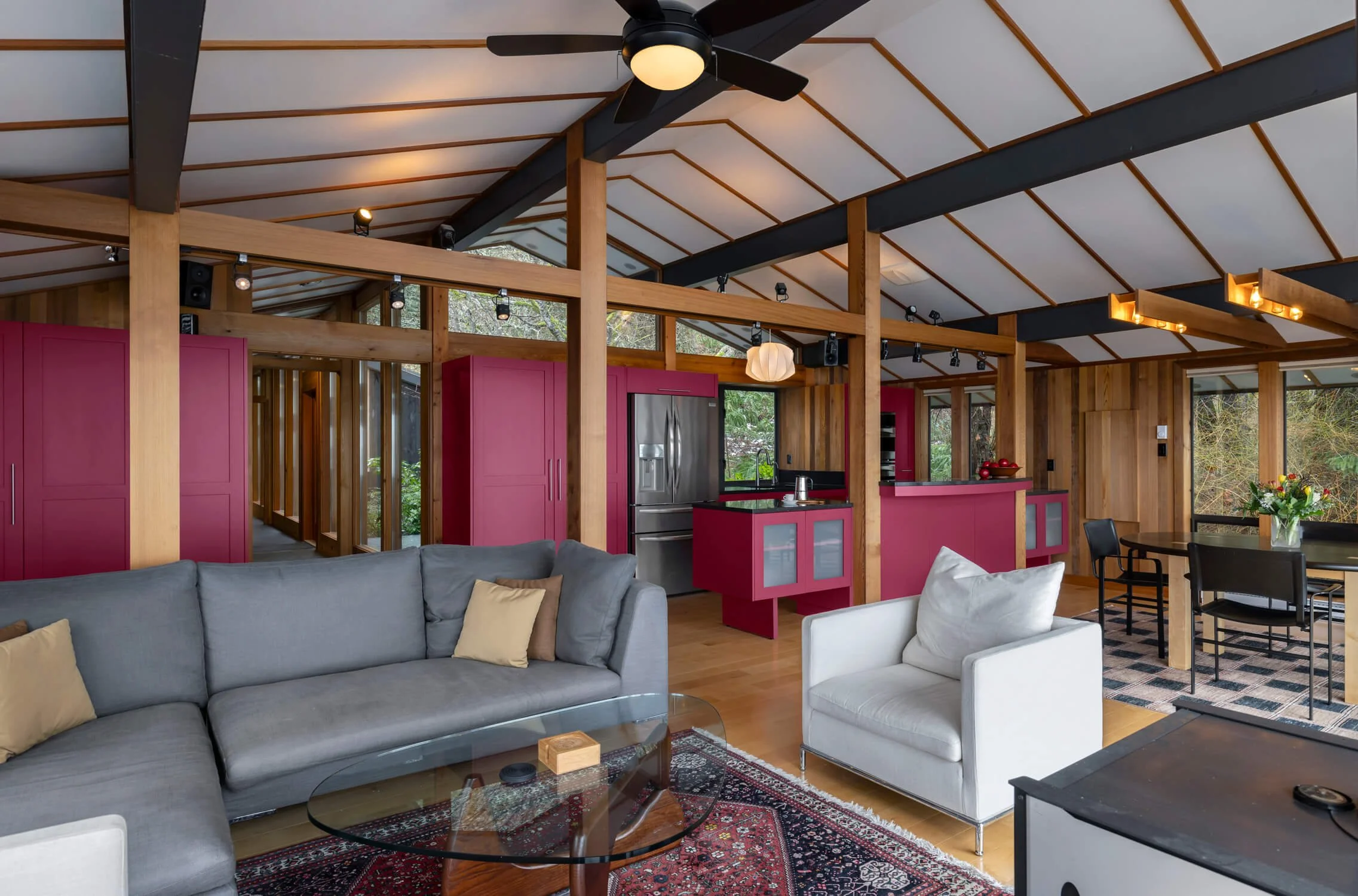 Living room and kitchen area with wood-paneled walls, pink cabinetry, a gray sofa with beige cushions, a white armchair, a glass coffee table, and a dining table with black chairs, illuminated by a ceiling fan and pendant lights.