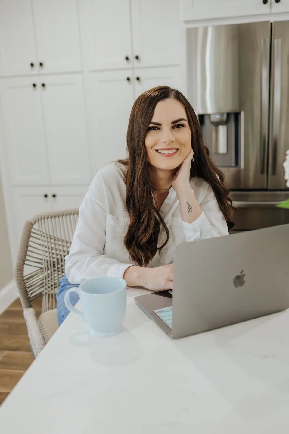 A woman with long brown hair and a tattoo on her wrist, sitting at a kitchen table with a laptop and a mug, smiling at the camera.