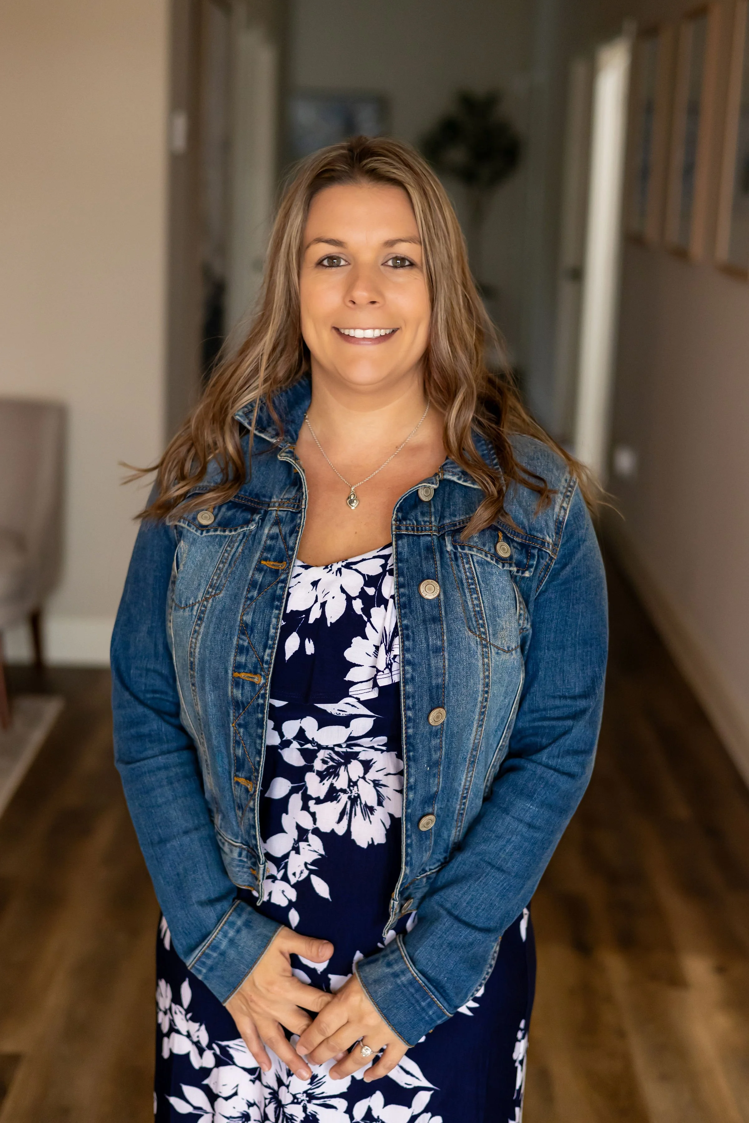 A woman with long brown hair smiling, wearing a denim jacket over a black and white floral dress standing inside a home.