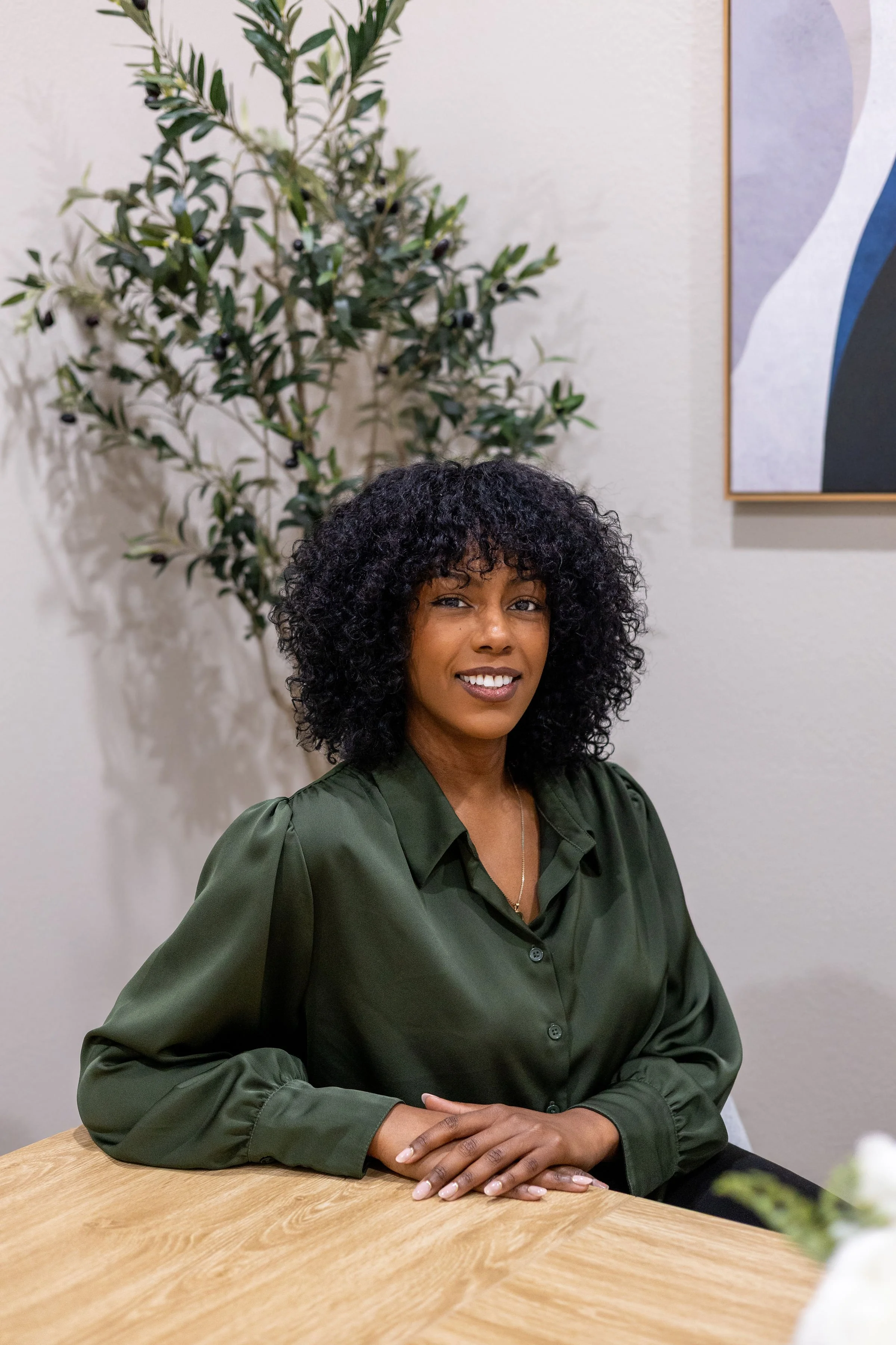 A woman with curly black hair and a dark green blouse sitting at a wooden table, smiling at the camera. A potted plant with dark berries and a framed abstract art piece are in the background.