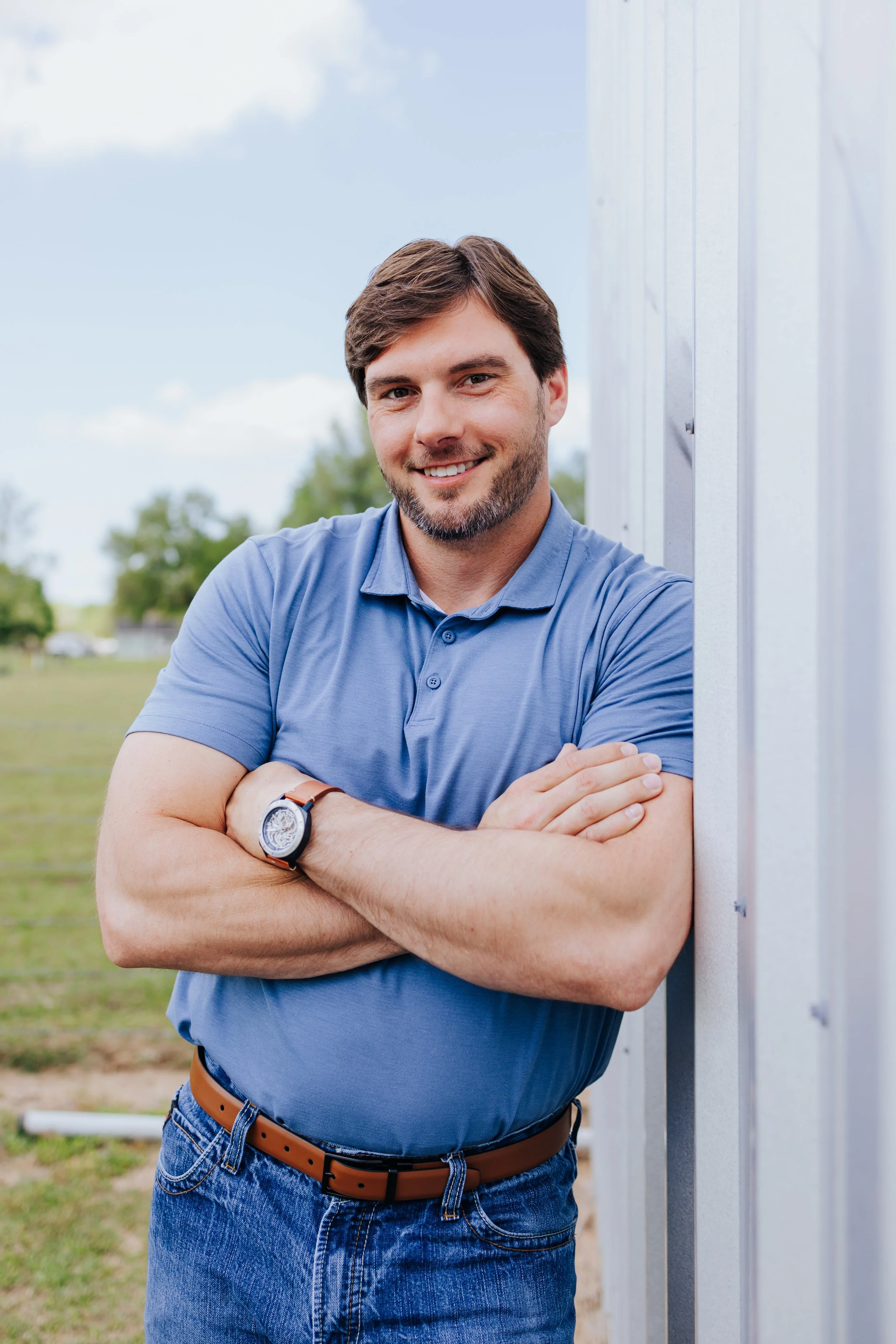 A smiling man with brown hair, beard, and blue eyes, wearing a blue polo shirt, standing outdoors with arms crossed near a metal structure, and trees and a grassy field in the background.