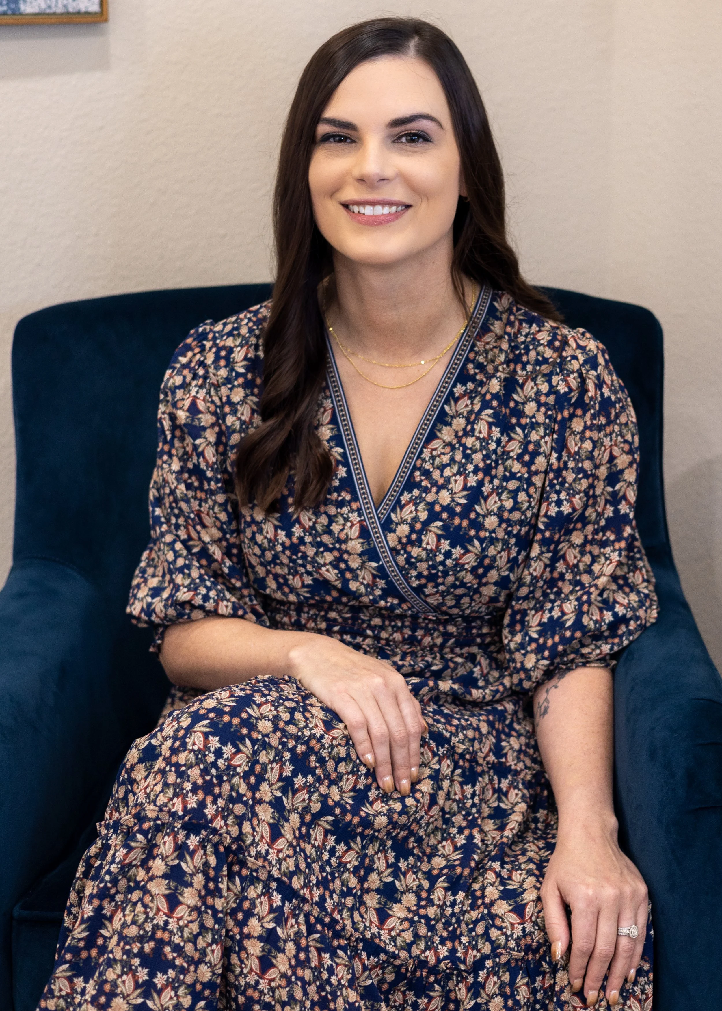 A woman with long dark brown hair, wearing a floral dress and gold necklaces, sitting in a dark blue armchair in front of a beige wall.
