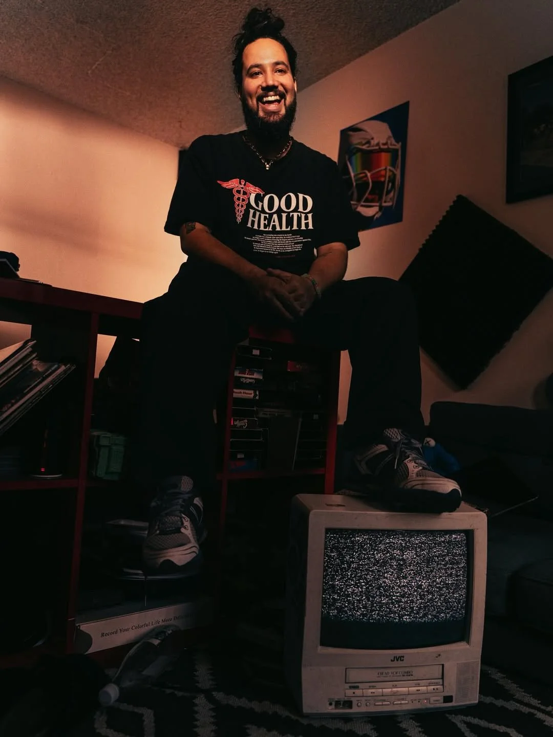 A man with a beard and long hair tied up in a bun is sitting on a red shelf in a room, smiling. He is wearing a black t-shirt with the words 'GOOD HEALTH' and a medical symbol, dark pants, and sneakers. There is a small, old television with static on the screen on the floor in front of him. The room has posters on the walls and various books and items on the shelves.