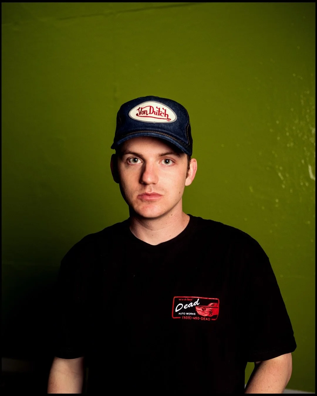 A young man with light skin, wearing a black t-shirt and a blue cap with 'Von Dutch' embroidered on it, stands against a green wall with water droplets.