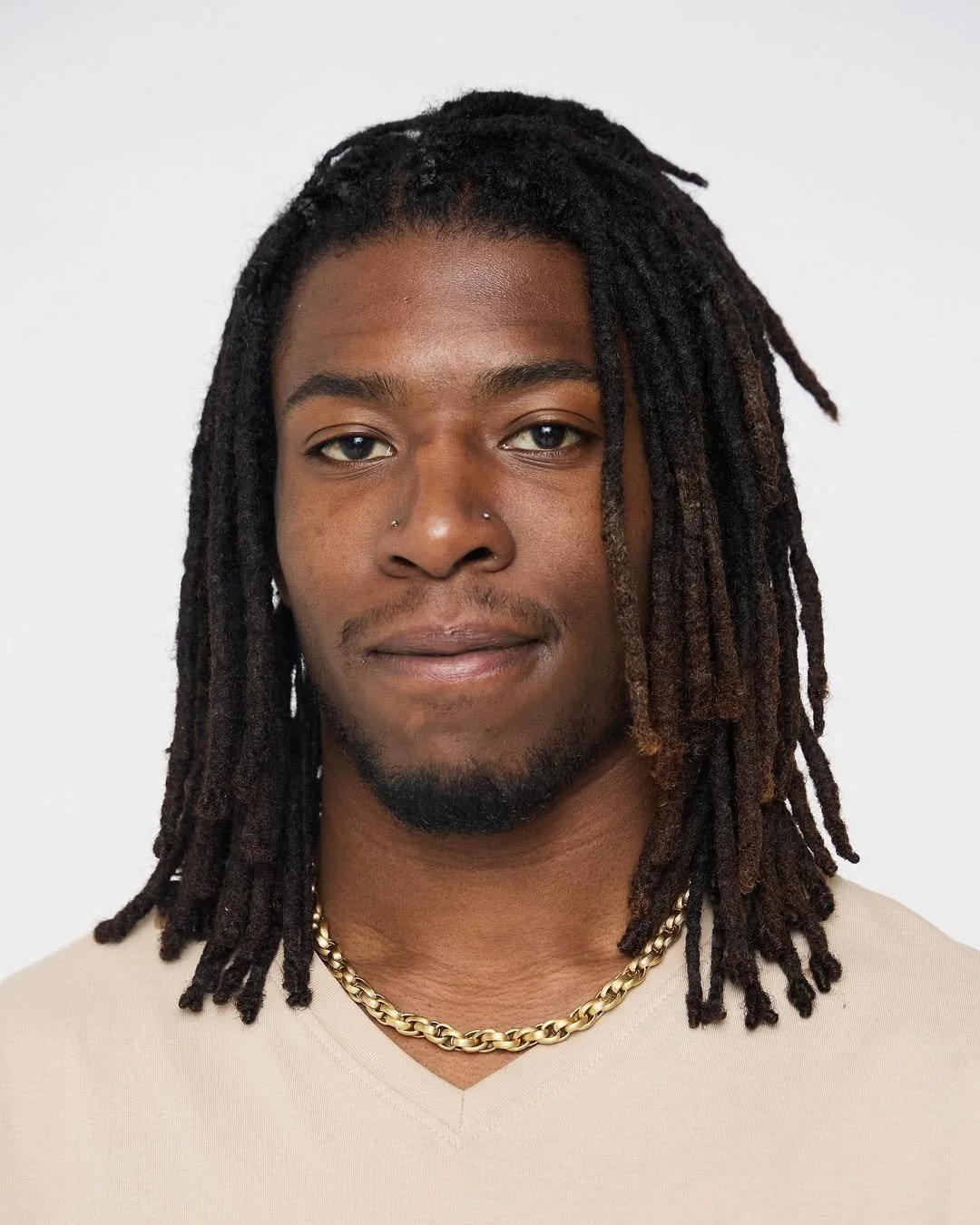 A young man with long dreadlocks, wearing a beige shirt and gold chain, looking directly at the camera against a plain white background.