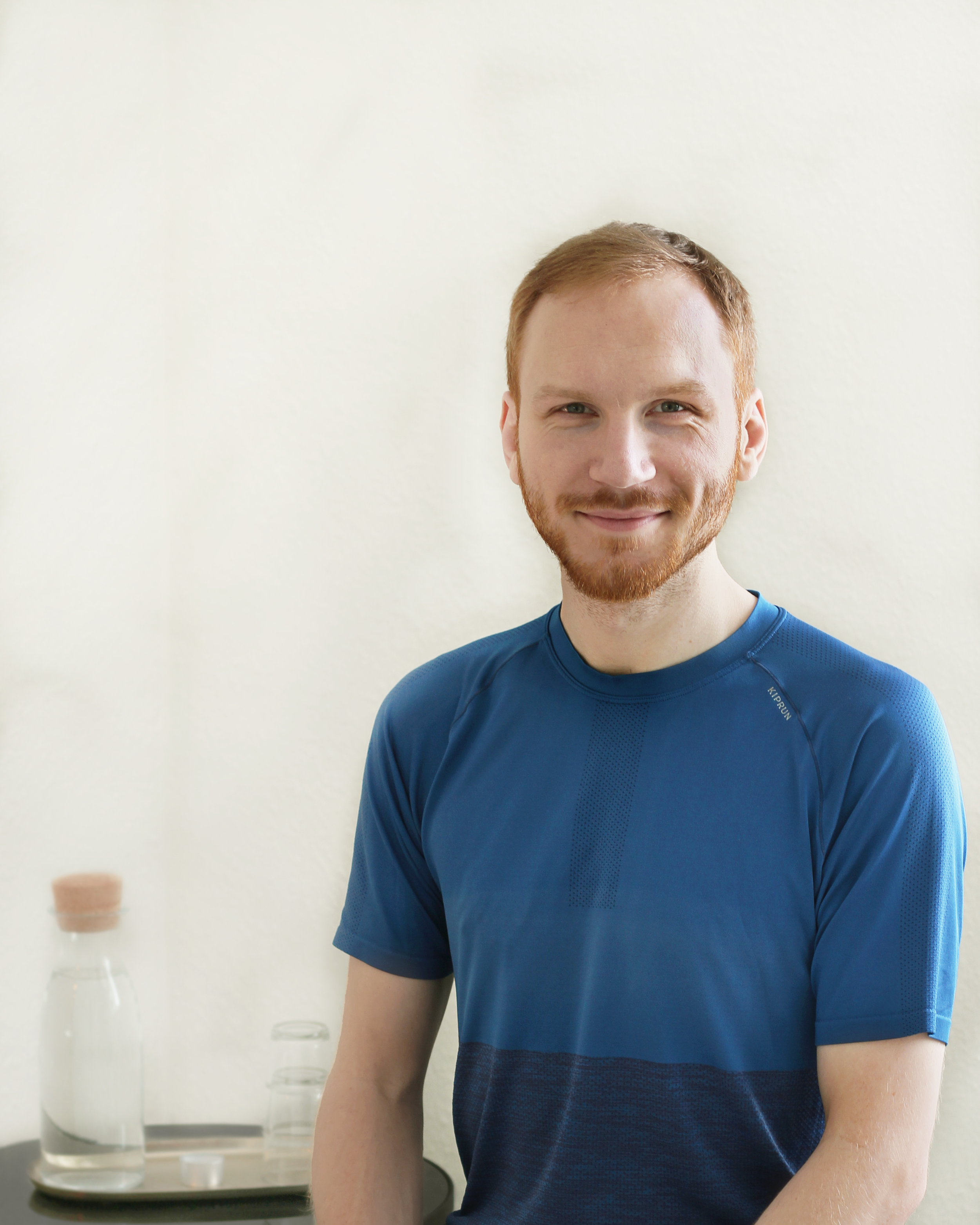 Lächelnder junger Mann mit roten Haaren und Bart trägt ein blaues Sportshirt und sitzt an einem Tisch, im Hintergrund Glaswasserflasche und Gläser auf einem Tablett.
