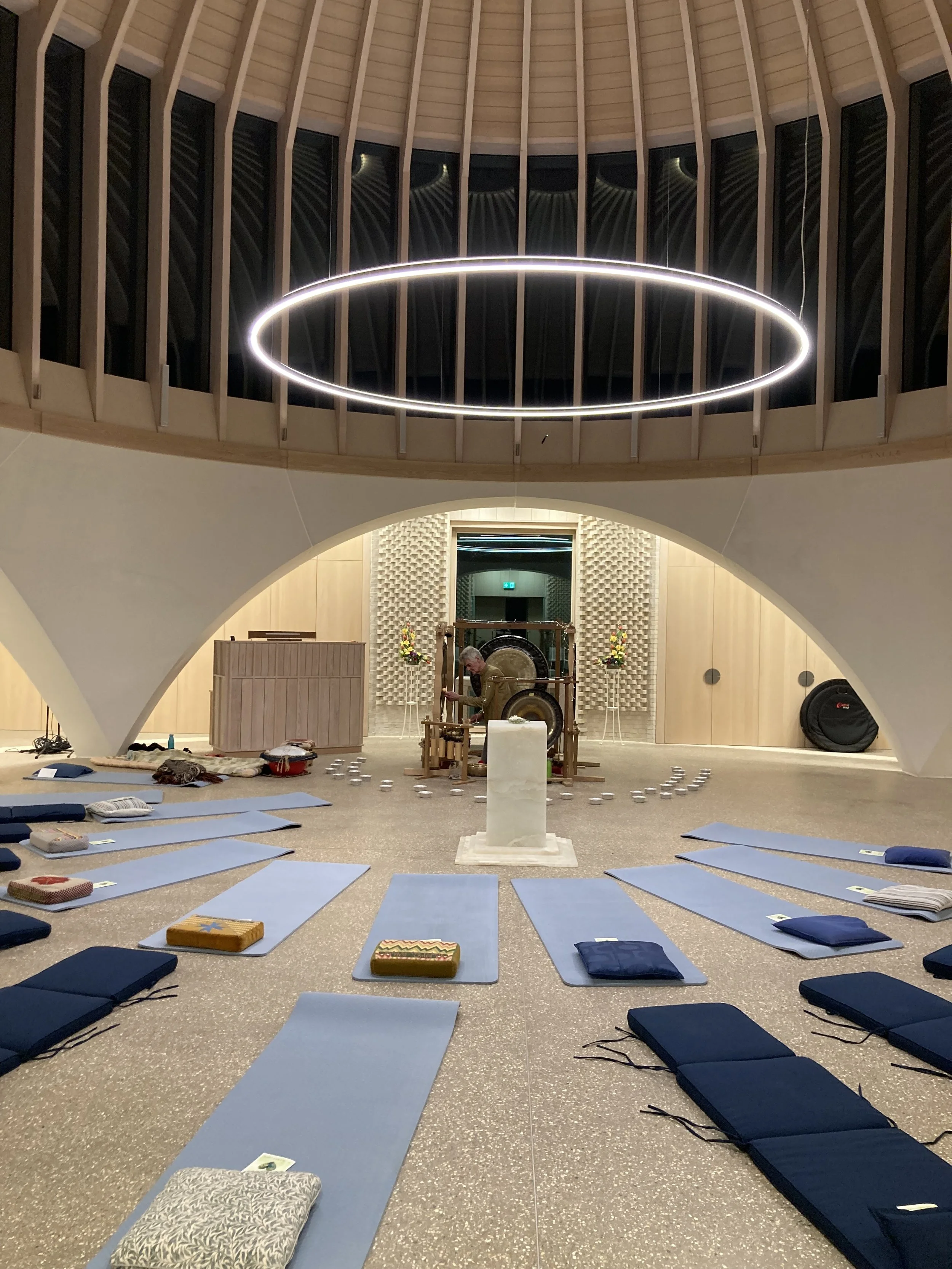 Indoor yoga or meditation space with blue mats and cushions arranged on the floor, a central statue, and a gong in the background, illuminated by a large circular ceiling light.