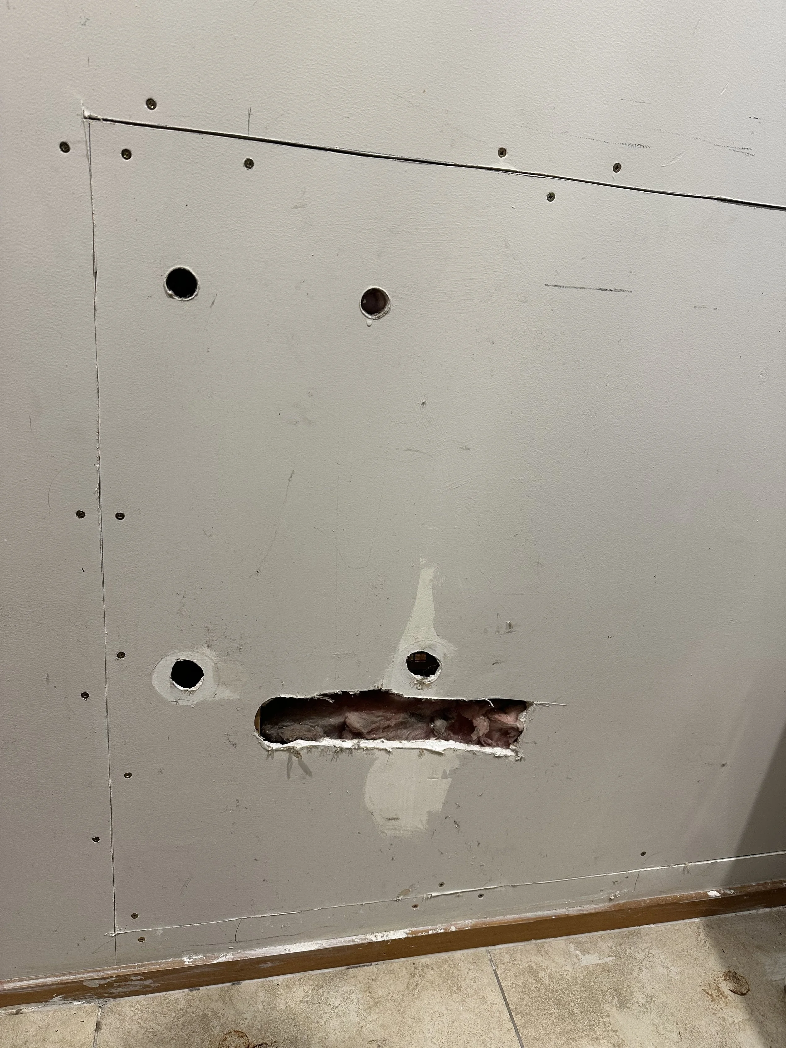 A drywall wall with multiple holes and a large opening at the bottom, exposing insulation and wiring behind the wall.