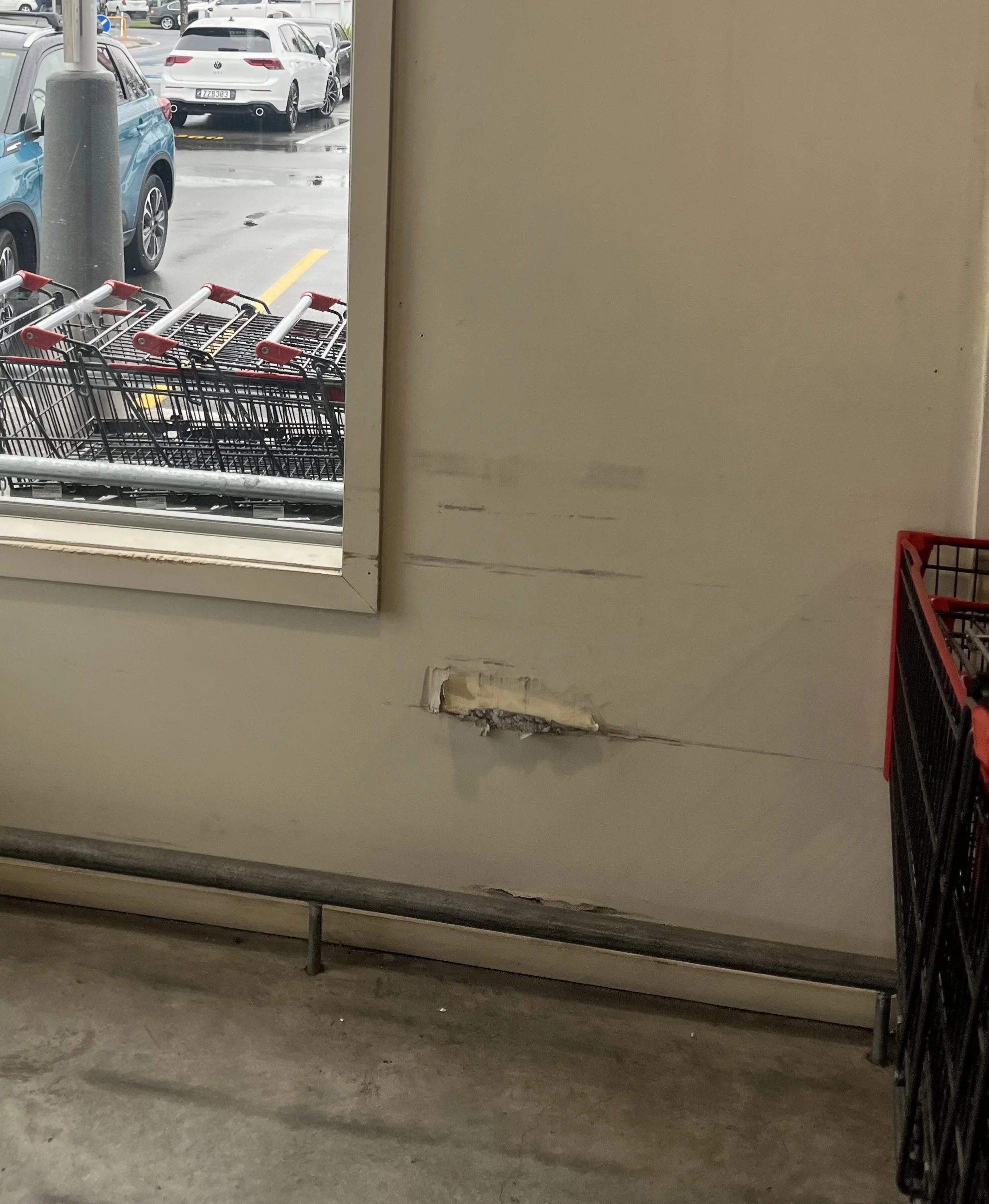View from inside a building looking out a window at a parking lot with cars. In the foreground, there are empty shopping carts stacked near the window. The floor is concrete, and the wall has a damaged area with some debris underneath.