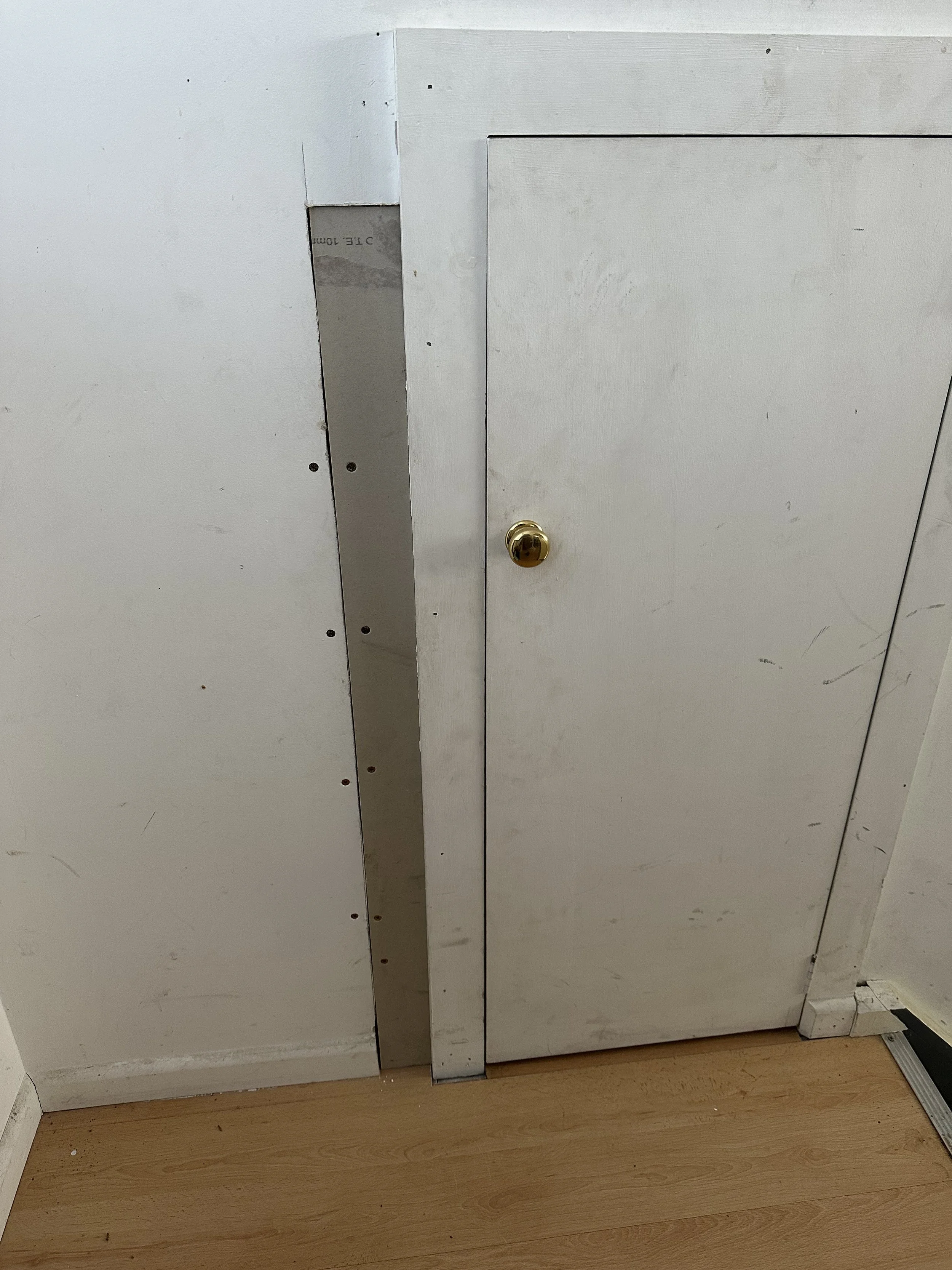 White cabinet or door with a round, golden handle, and a vertical strip of metallic material in the center. The cabinet has visible scratches and marks, and the bottom edge shows a slight gap and scuff marks. The floor is wooden with some dirt and debris.