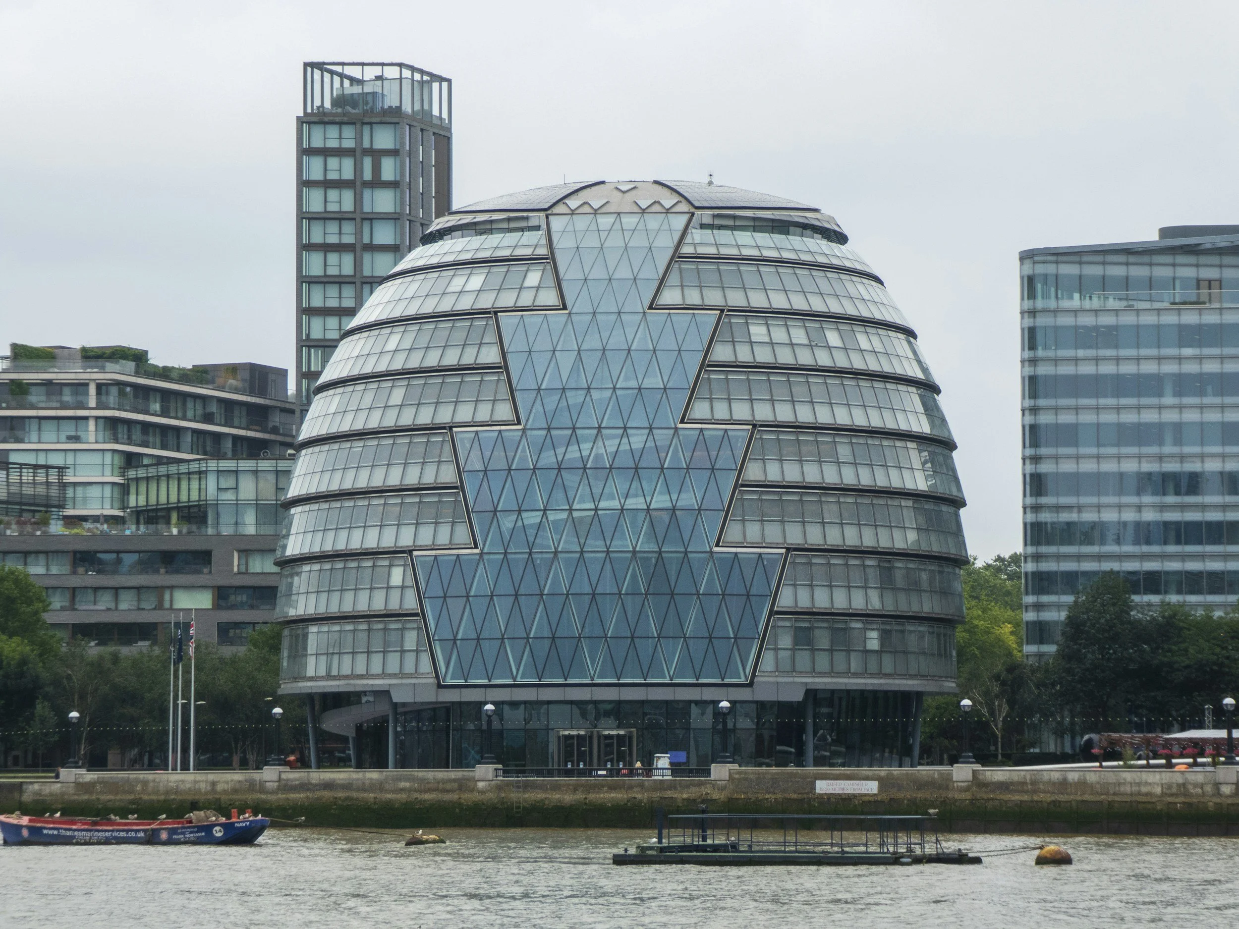A modern building with a rounded, glass exterior and geometric patterns, with taller buildings nearby, situated along a river with a boat passing in the foreground.