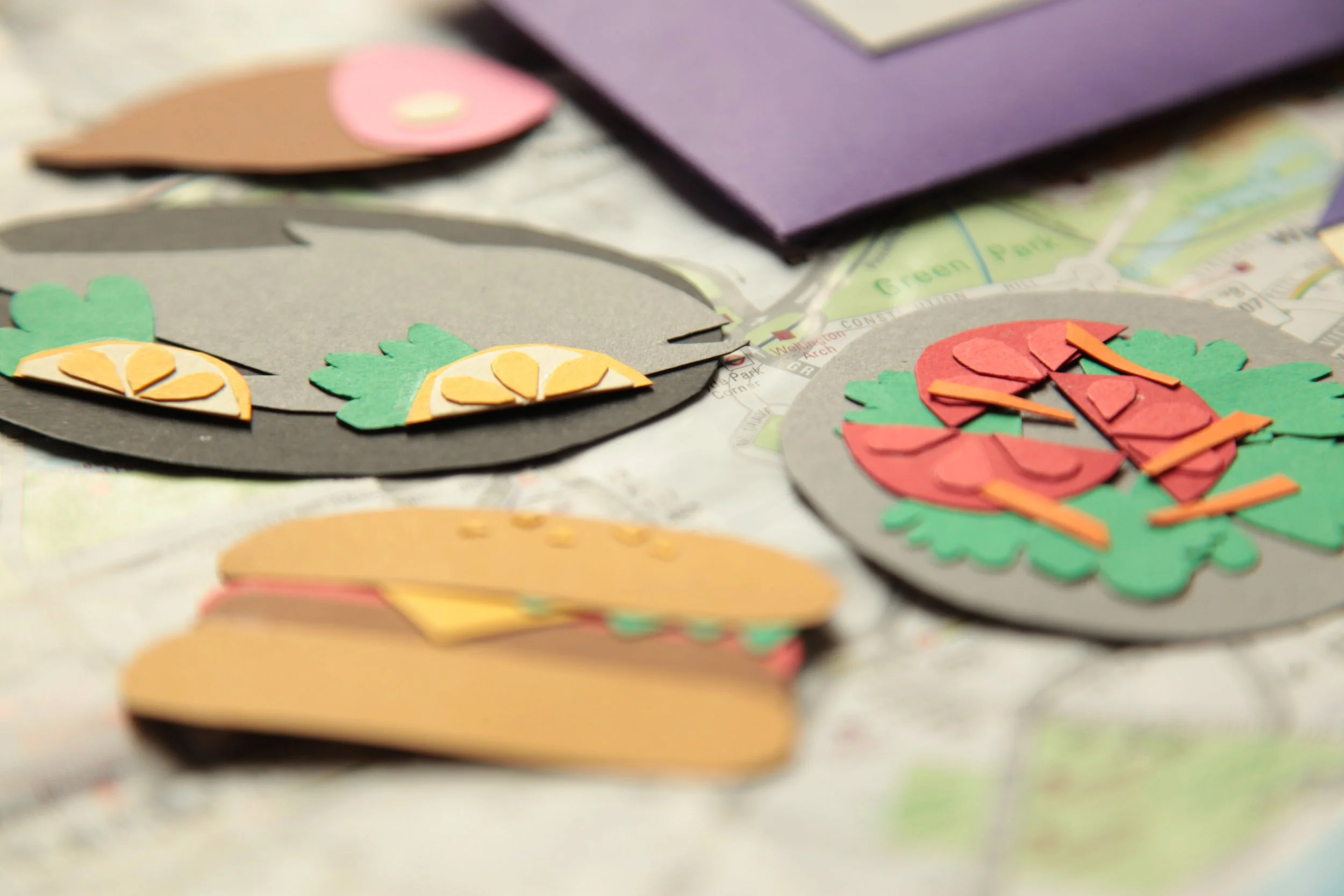 Colorful paper cutouts of fruit slices, including lemons, strawberries, and peaches, arranged on a table with a map and purple paper in the background.