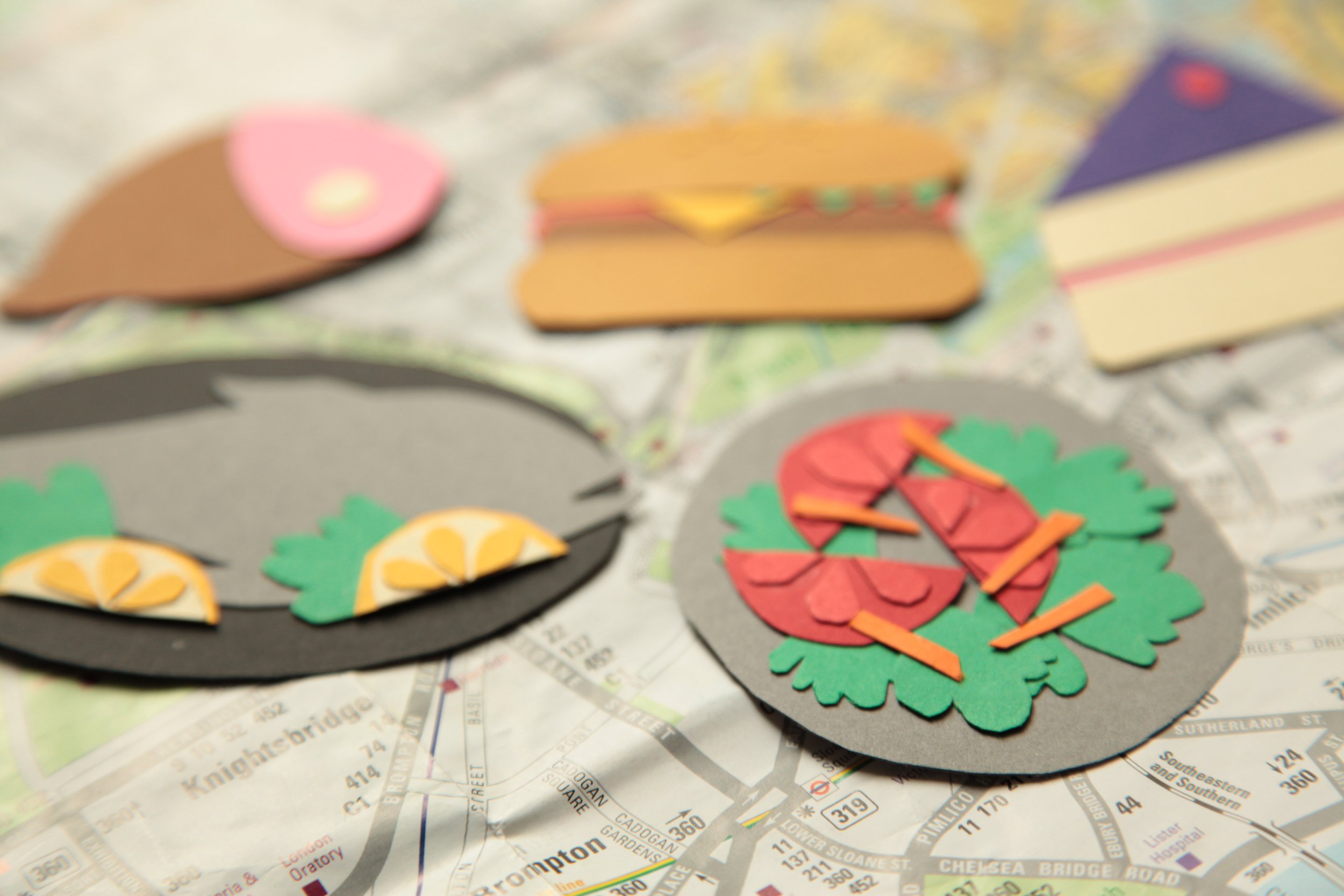 Colorful paper food cutouts of a burger, tacos, a taco salad, and a bowl of chopped vegetables placed on a city map.