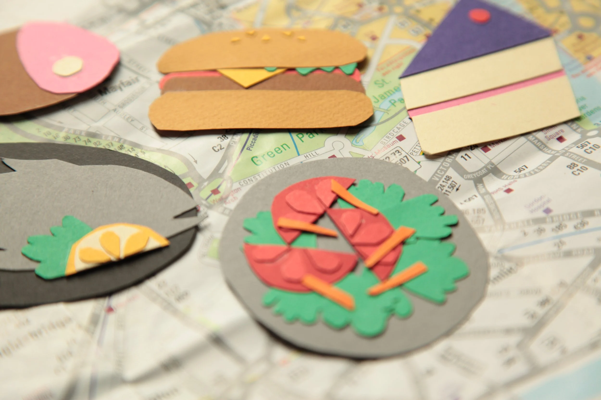 Craft paper food and picnic items placed on a map, including a pizza, a sandwich, and a drink with a lemon wedge.
