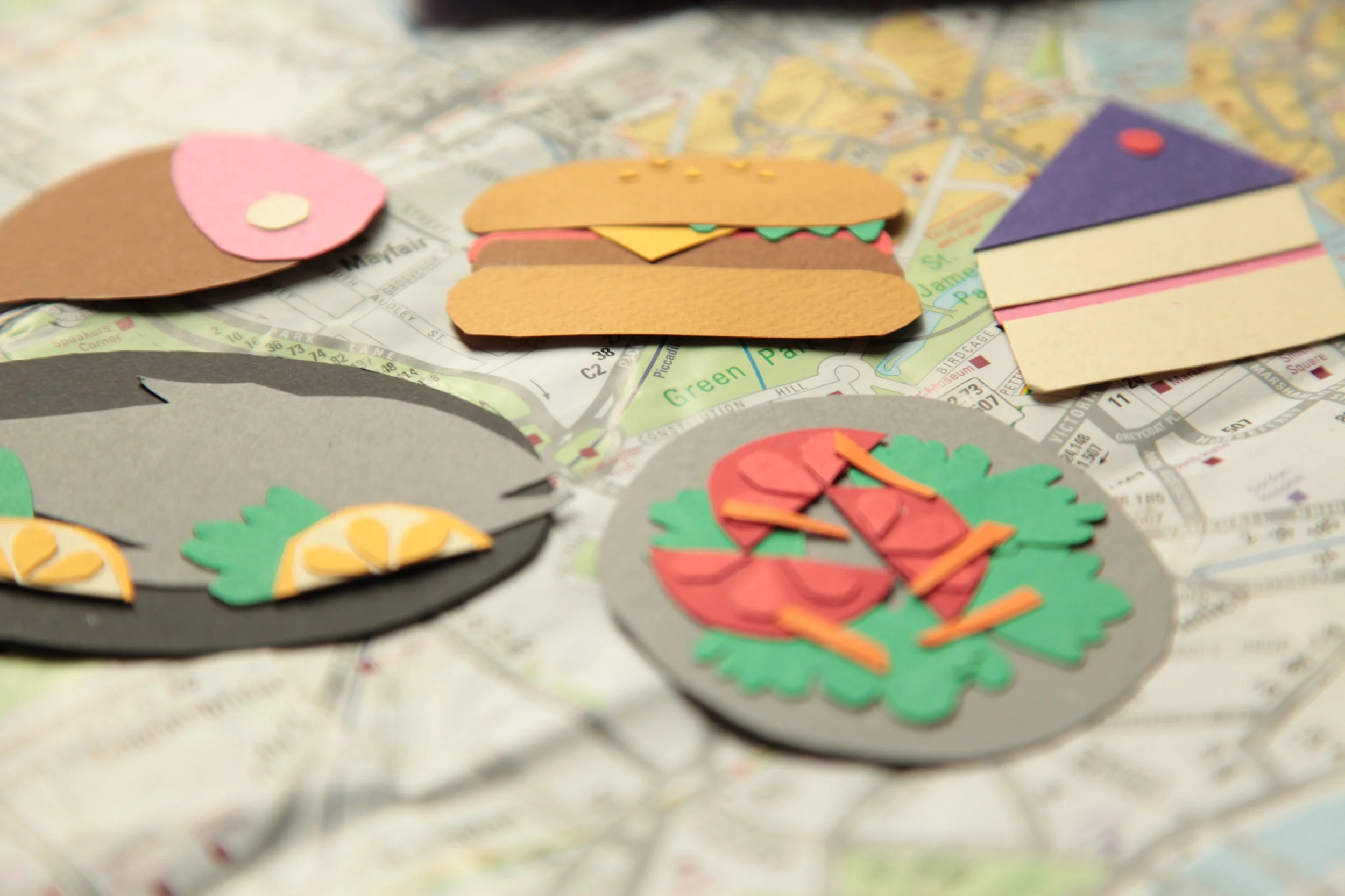 Colorful paper cutouts of food items like a taco, a burger, a pizza slice, and sushi, placed on a city map.