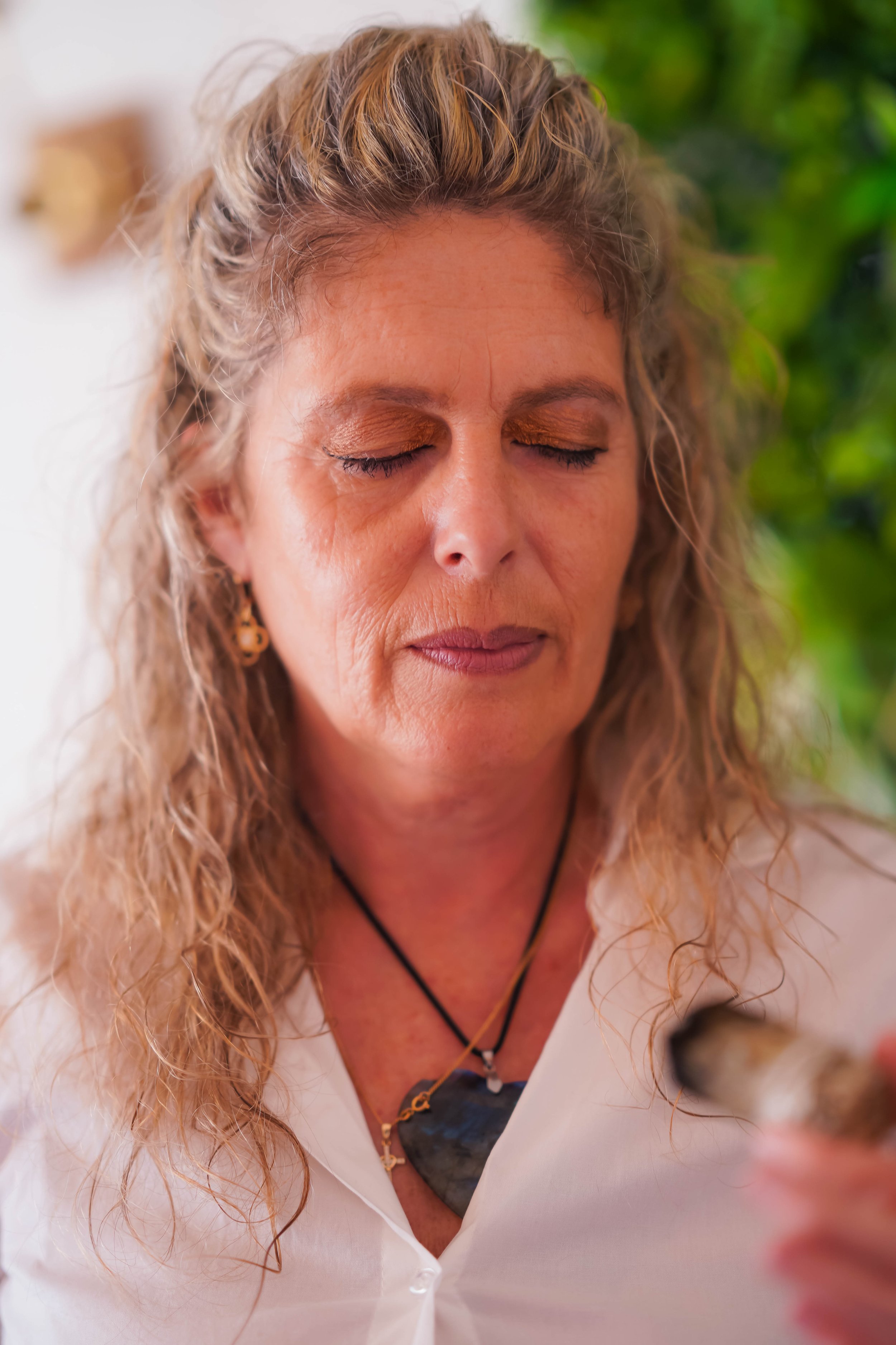 Femme aux cheveux blonds longs tenant un objet dans une main, portant un collier avec un pendentif en pierre, les yeux fermés, sur un fond flou de verdure.