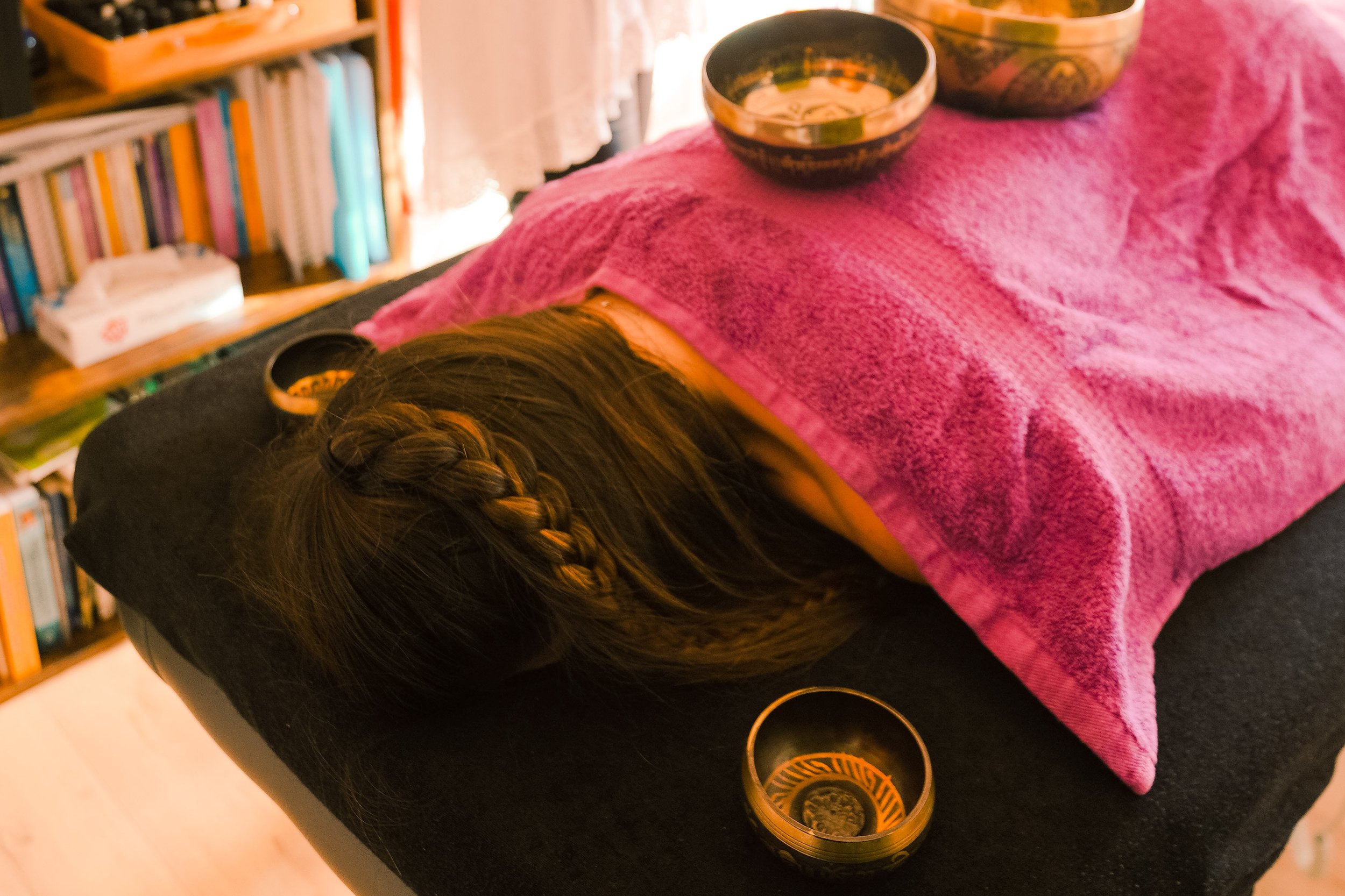 Personne allongée sur une table de massage couverte par une serviette rose avec des bols chantants tibétains placés sur le dos.